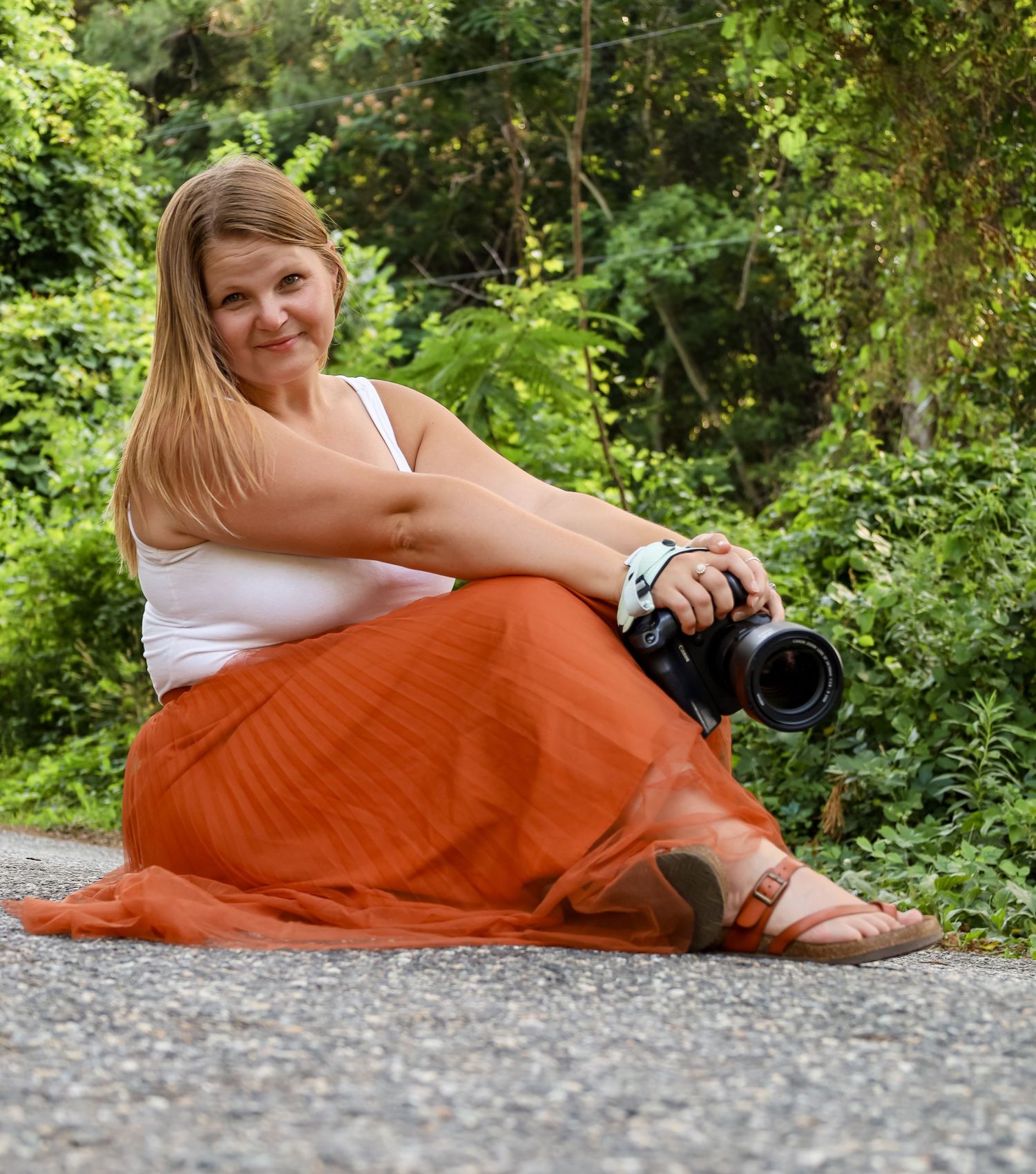 Woman in orange skirt sitting on pavement holding a camera, smiling outdoors with green trees behind her.