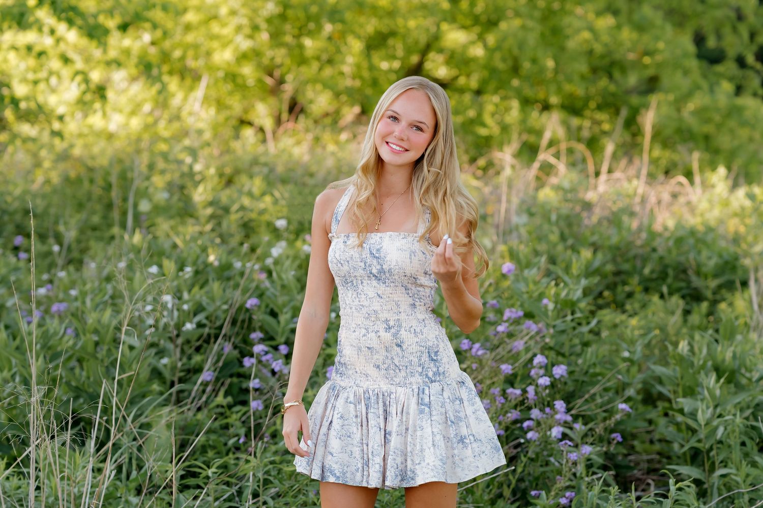Young blonde woman in floral sundress smiling outdoors surrounded by lush green wildflowers and purple blooms.