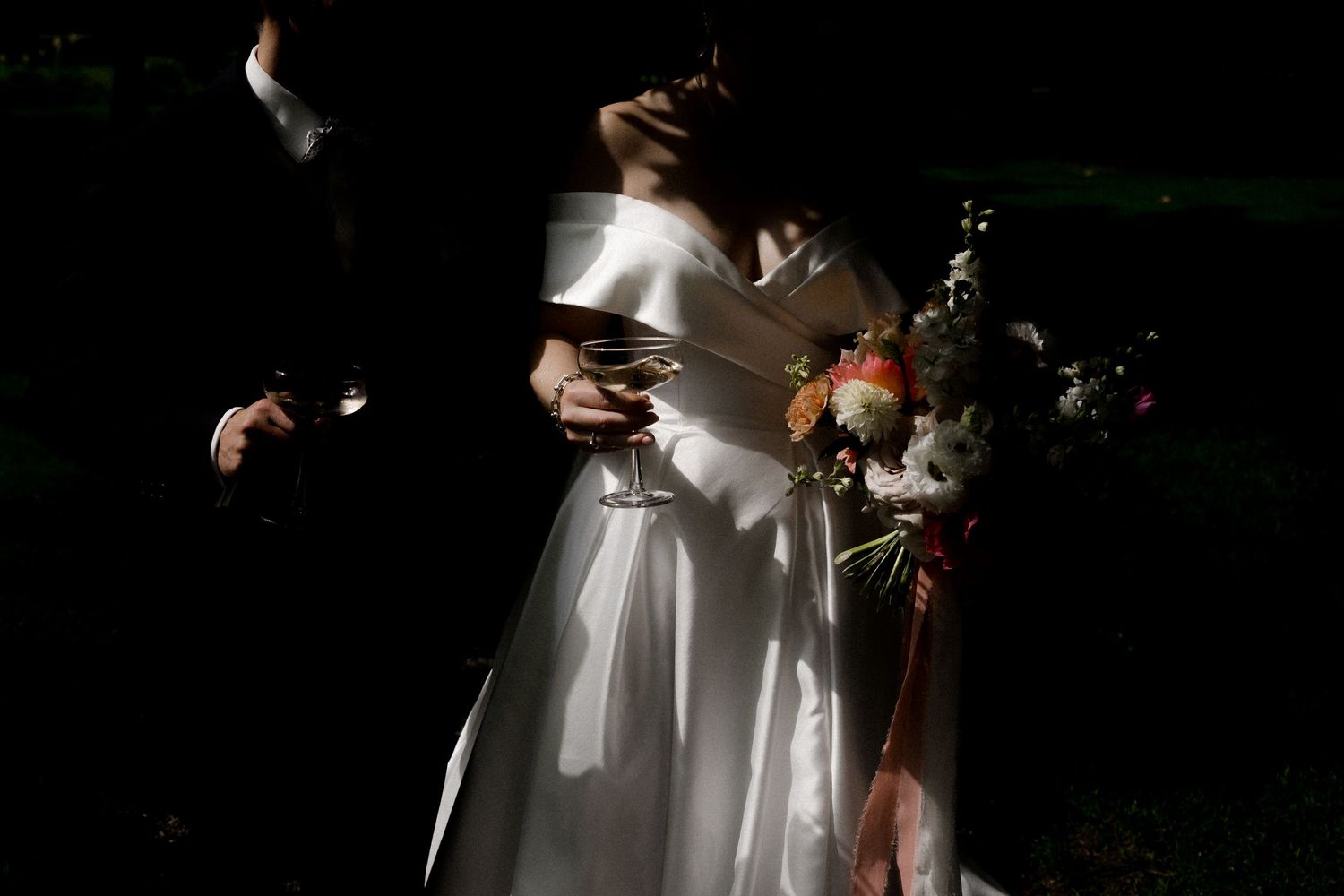Bride in white gown holding bouquet of flowers and wine glass with groom in black suit at wedding ceremony.
