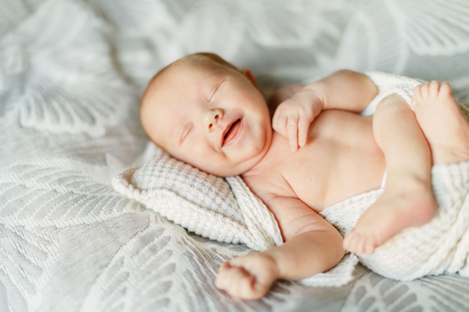 Peaceful newborn baby smiling while sleeping on a soft pillow, wrapped in a white blanket on a cozy bed.