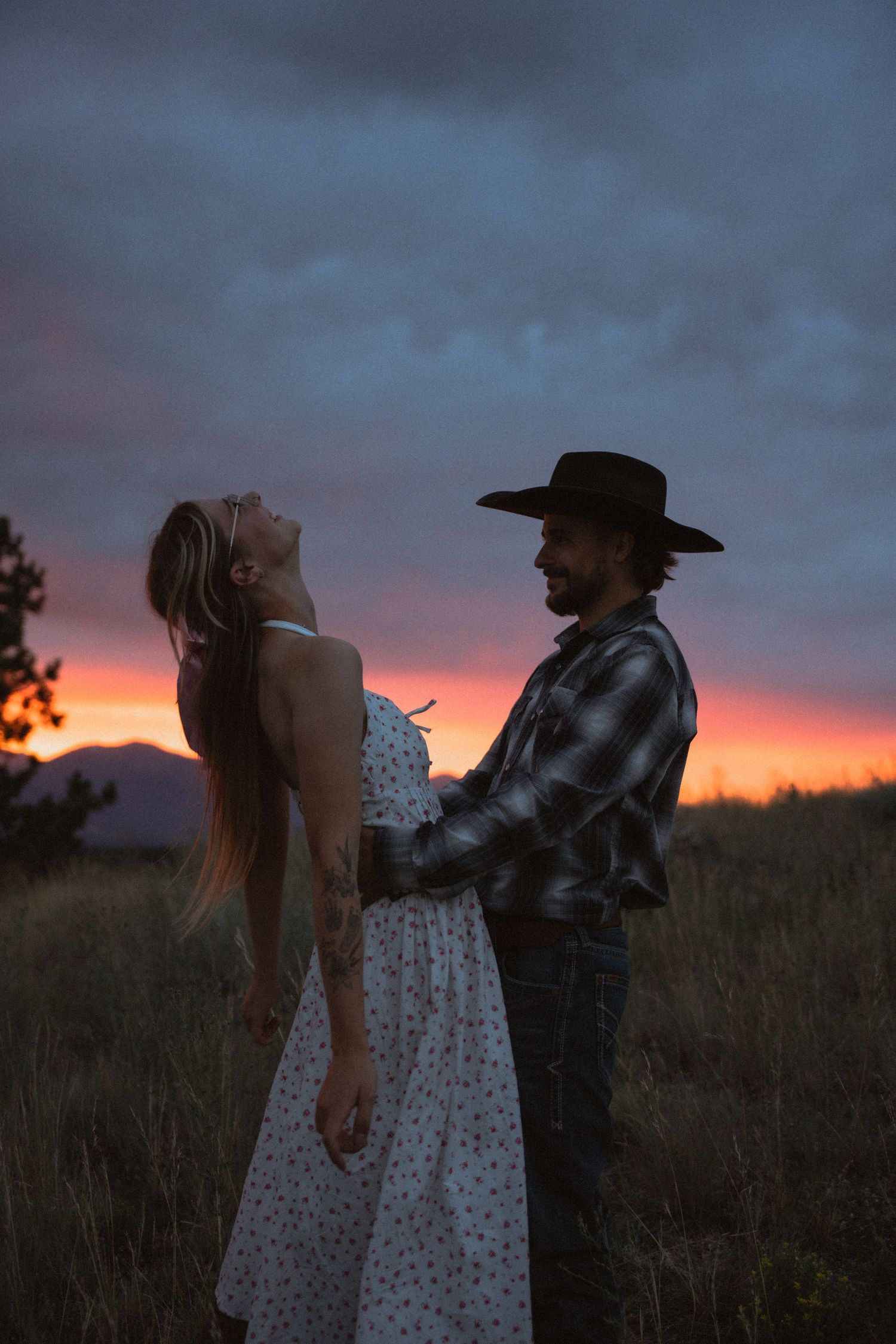 A couple embraces in a field at sunset, man in cowboy hat and plaid shirt, woman in floral dress, dramatic sky.