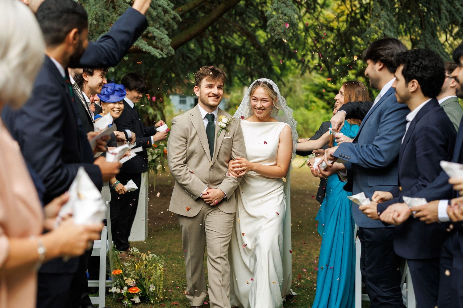 Newlywed couple walks down outdoor wedding aisle as guests toss confetti in a garden ceremony.