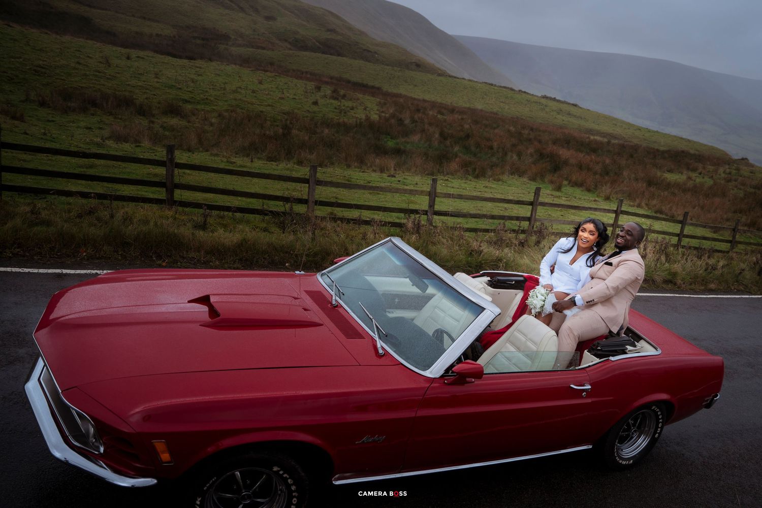 Couple posing in a red classic Ford Mustang convertible on a scenic road with green hills backdrop.