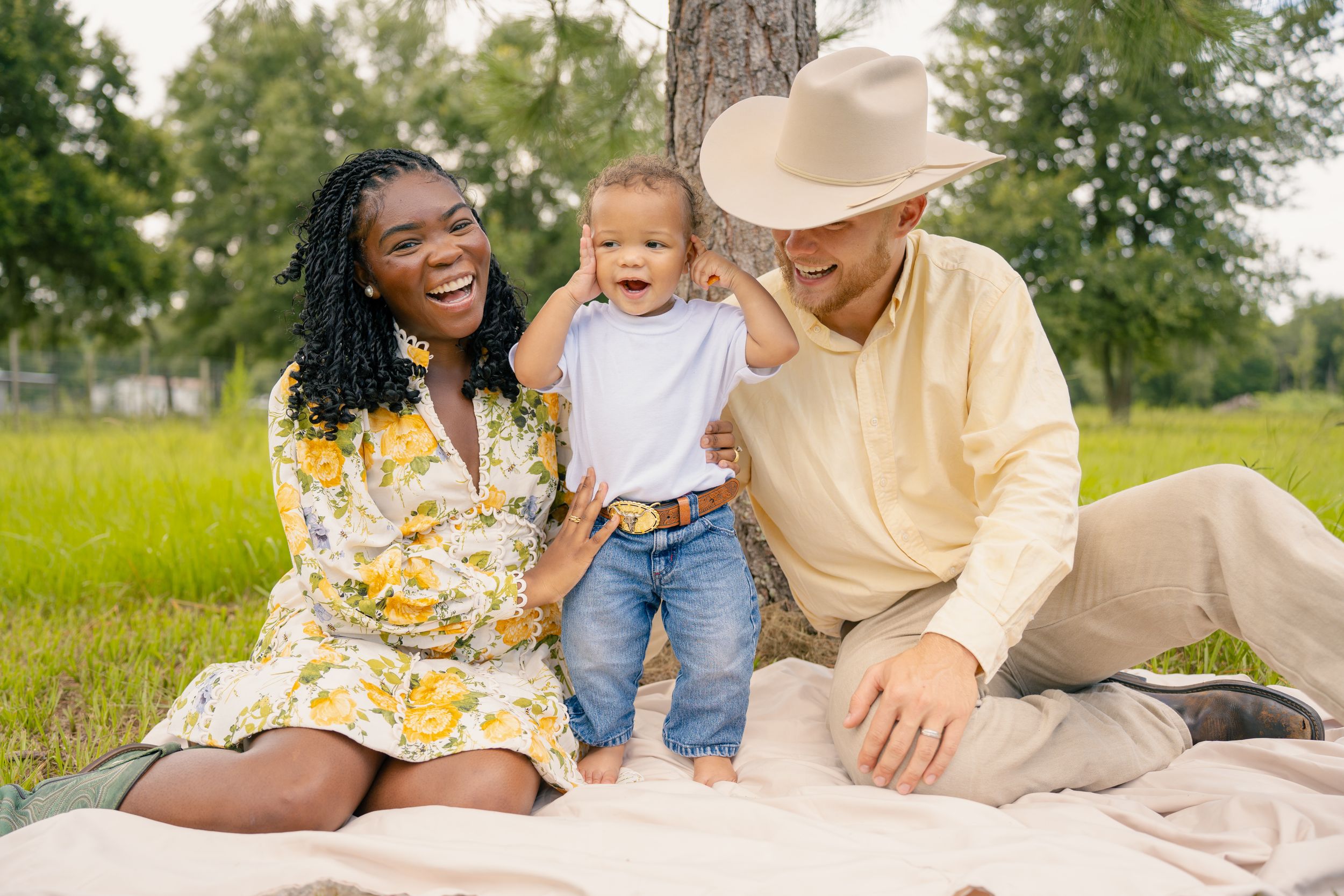 Happy interracial family with toddler sitting on blanket outdoors, laughing together in a green park setting.