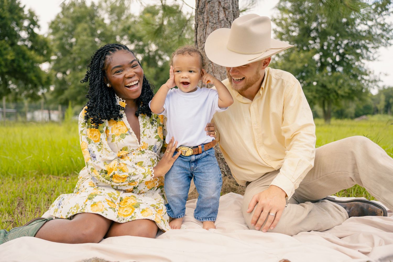 Happy interracial family with toddler sitting on blanket outdoors, laughing together in a green park setting.