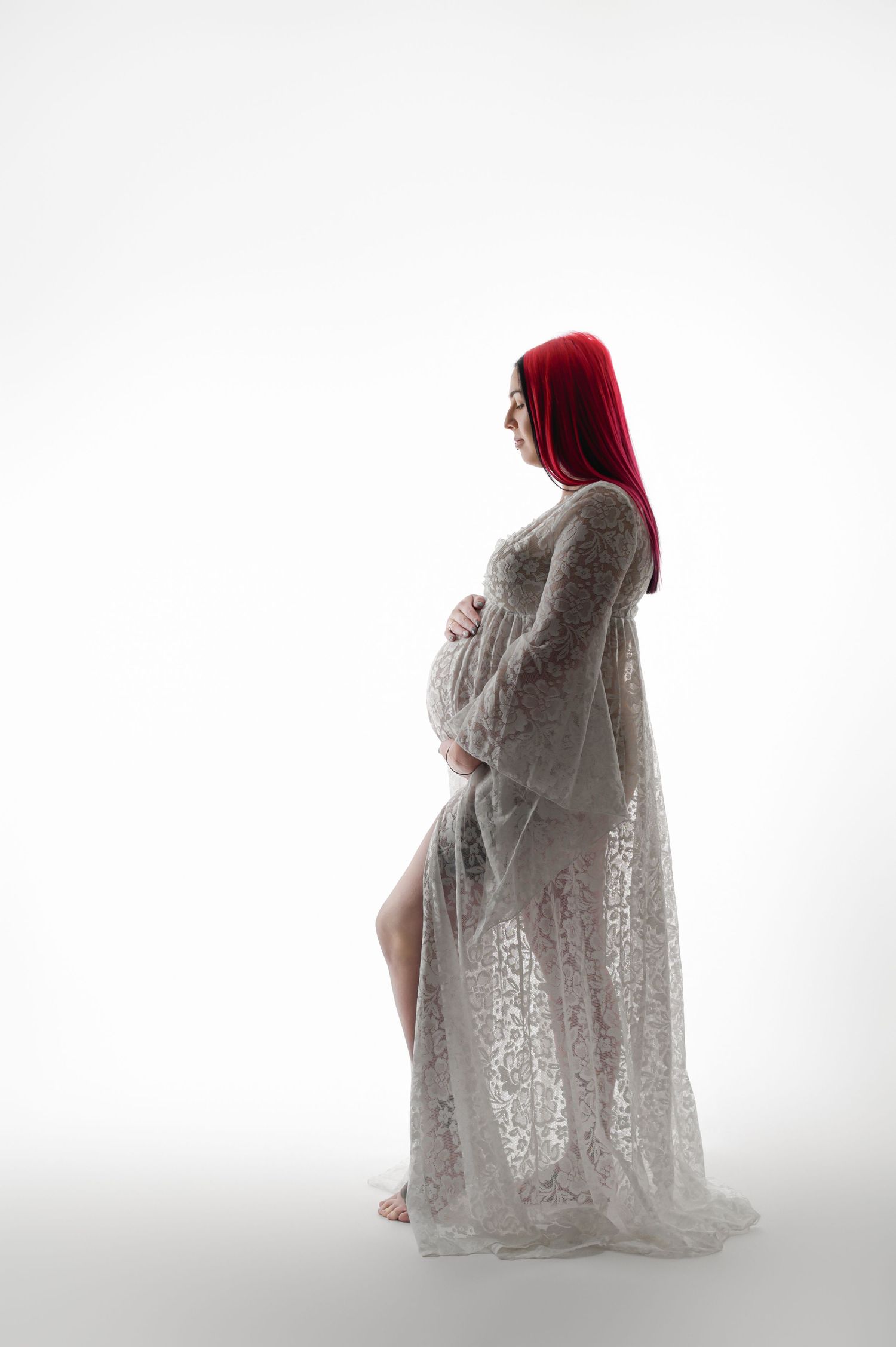 Pregnant woman with red hair wearing a white lace maternity gown, cradling her baby bump in a studio.