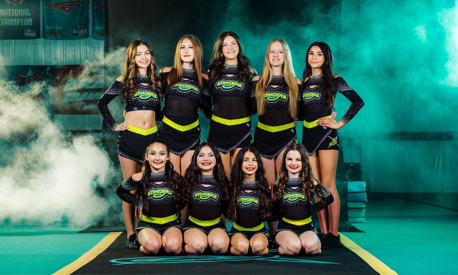 Cheerleading squad in black and neon yellow uniforms posing in two rows with dramatic teal smoke backdrop.