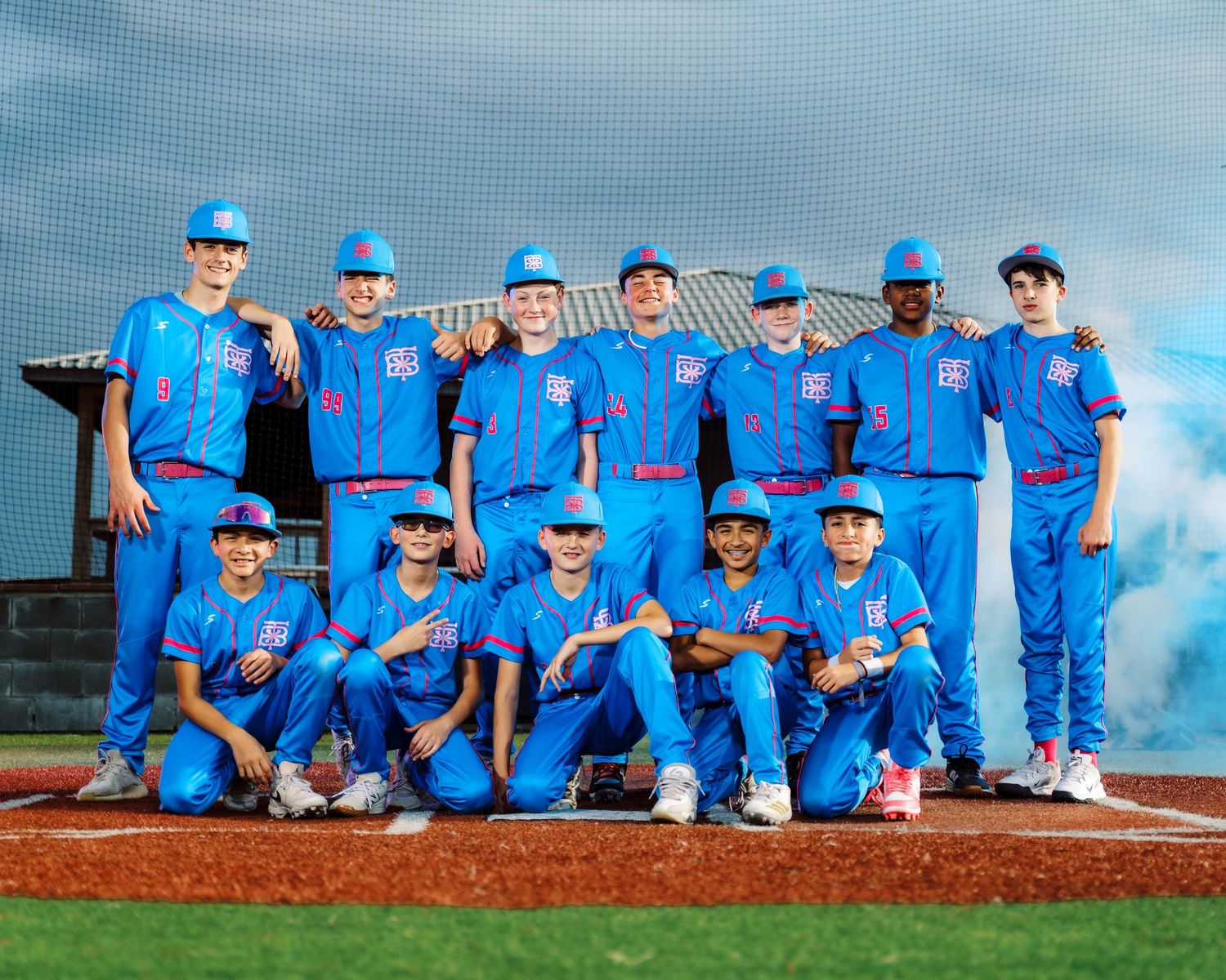 Youth baseball team posing in blue uniforms on a baseball field with colorful smoke in the background.