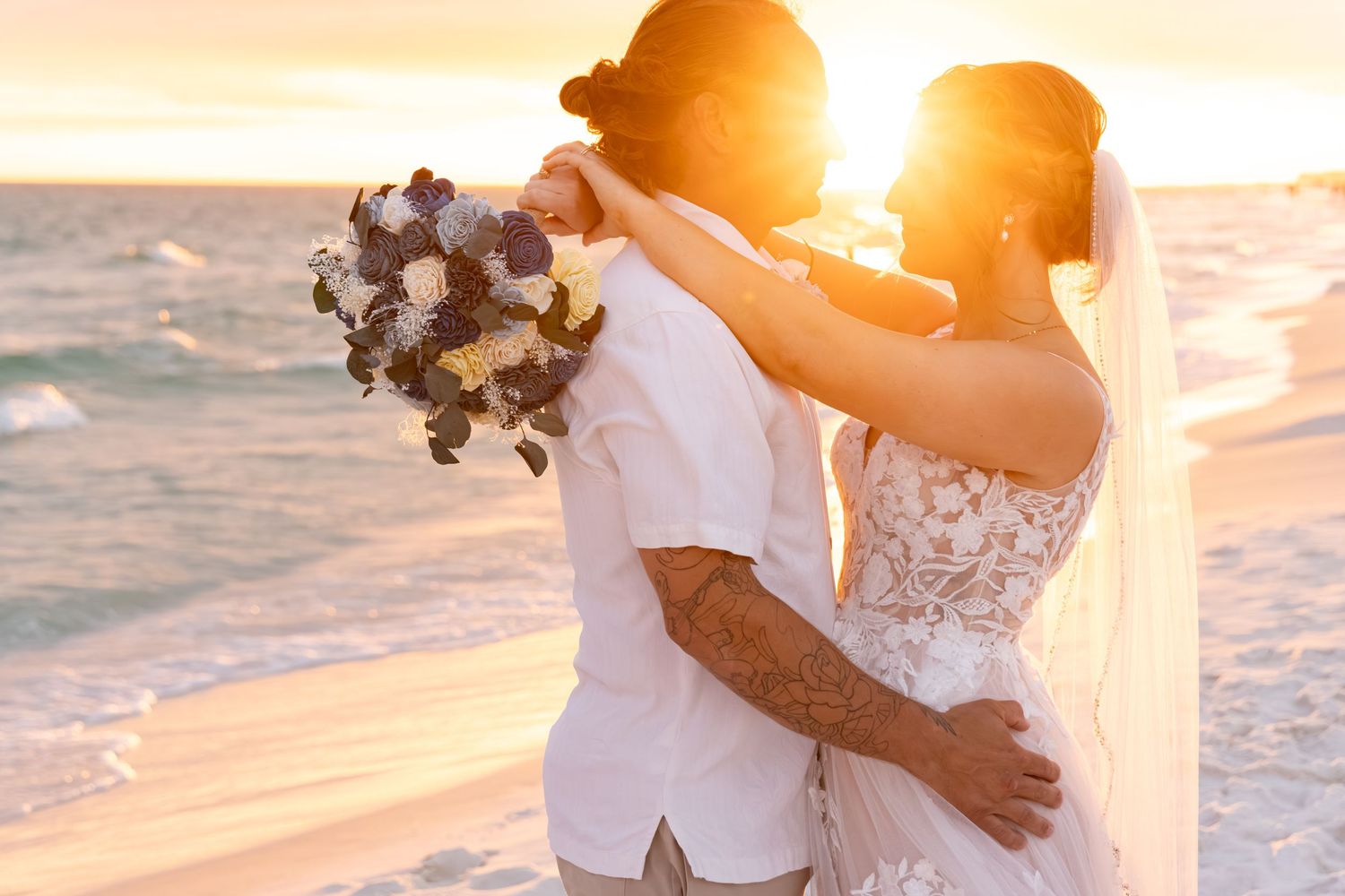 Newlywed couple sharing a romantic sunset beach wedding moment, bride in lace gown with veil holding floral bouquet.