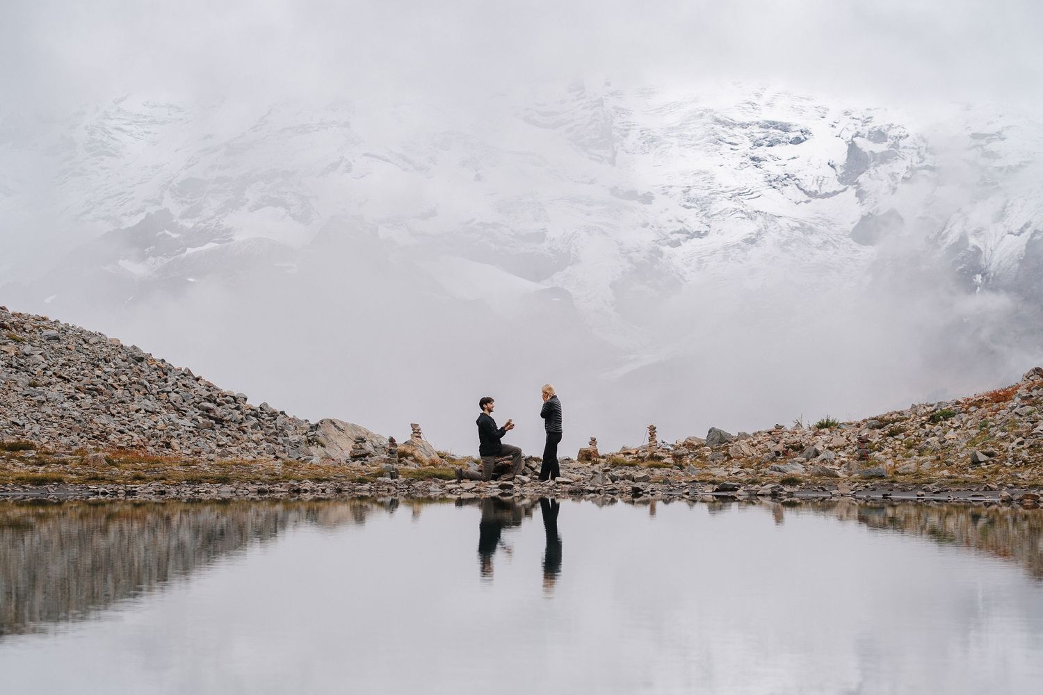 A couple shares a romantic proposal moment beside a reflective alpine lake with snow-capped mountains in the background.