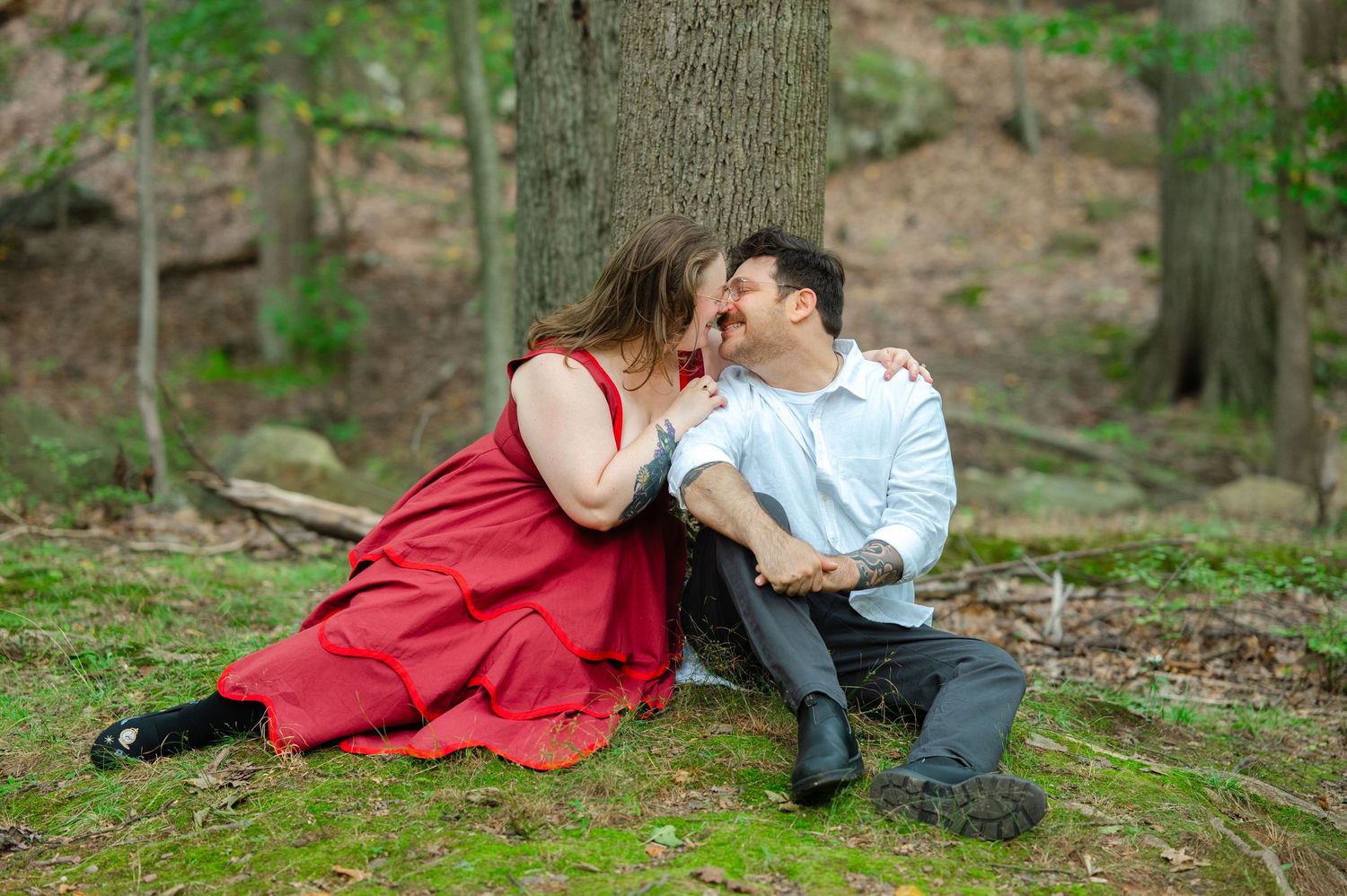 Couple cuddling and giggling during an engagement session in the woods in NJ. Woman wears a red dress and the man wears a white shirt.