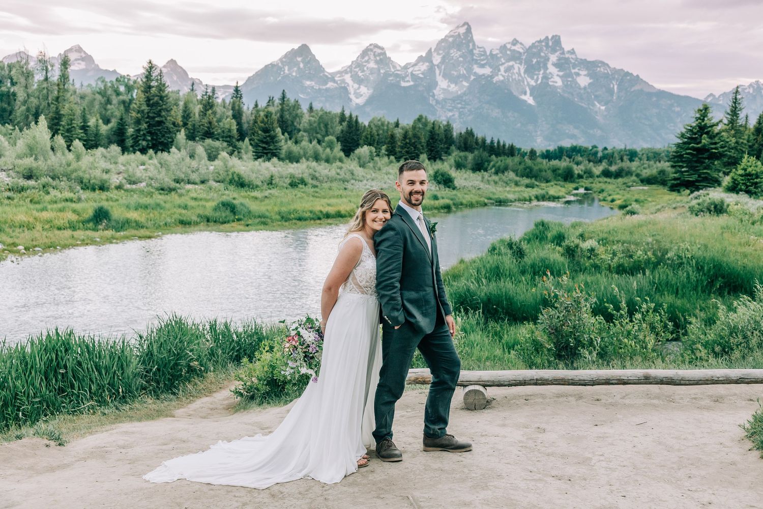 Bride in a white gown and groom in a dark suit pose together beside a calm river with snowy mountain peaks at Schwabacher's Landing.