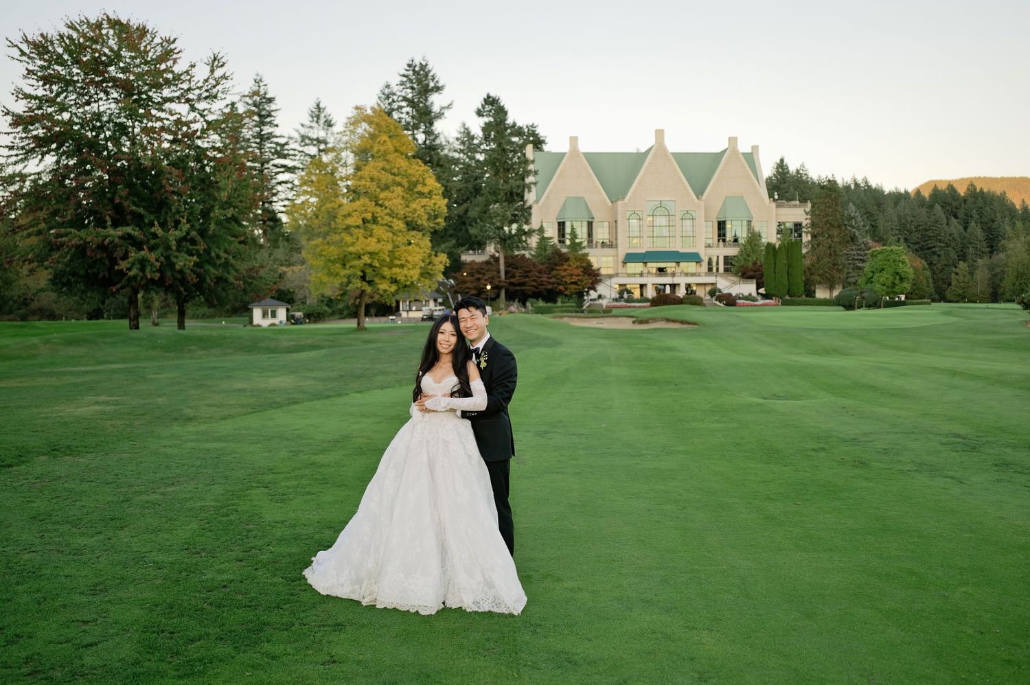 Bride in a flowing white gown and groom in a black tuxedo embrace on a lush green golf course lawn with a grand estate in the background.