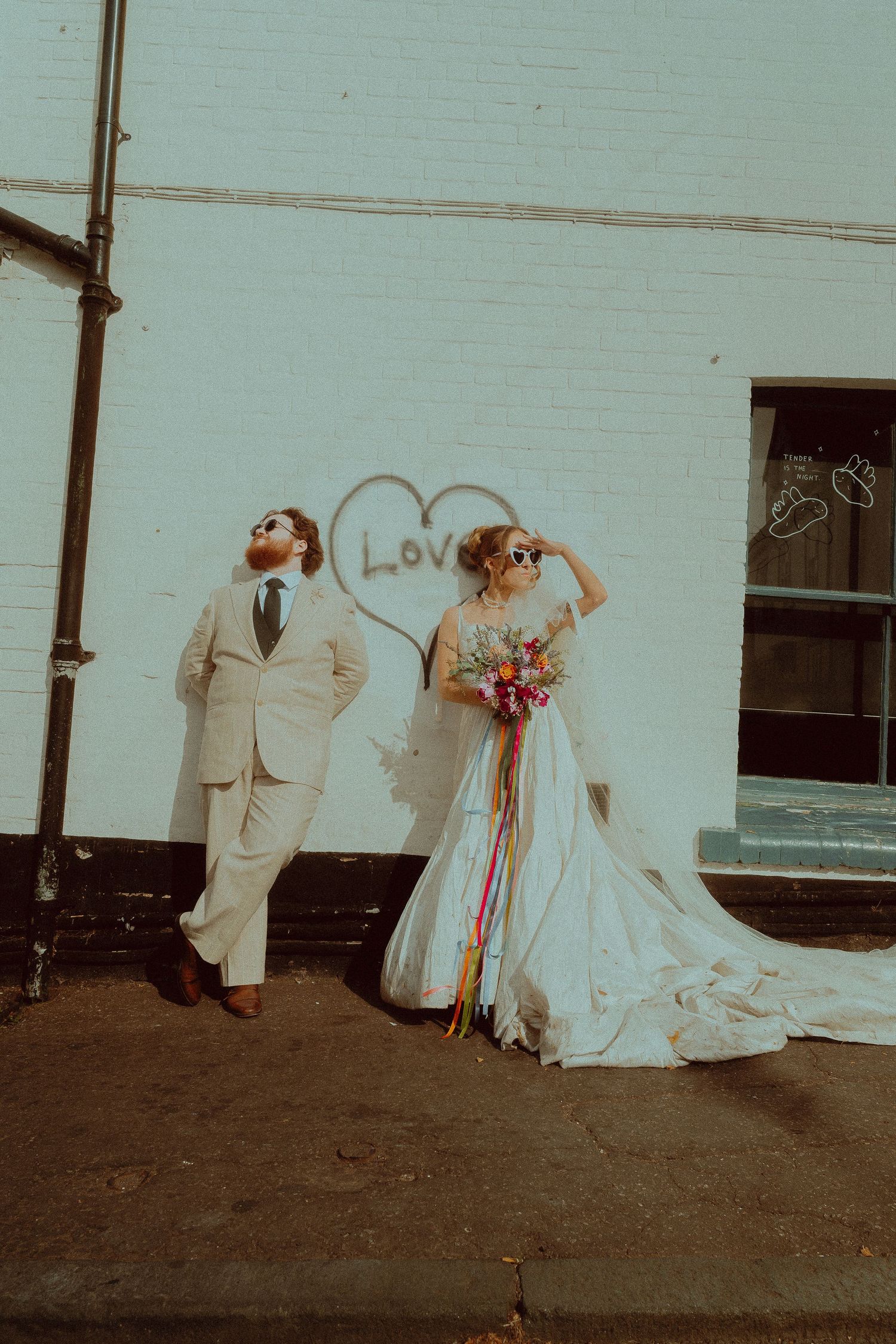 Bride in a flowing white gown with a colorful bouquet and groom in a beige suit pose against a white brick wall with a heart graffiti.