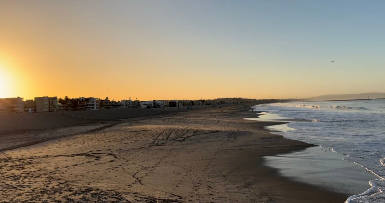 Golden sunset over a serene beach with gentle waves, sandy shore, and coastal buildings in the distance.