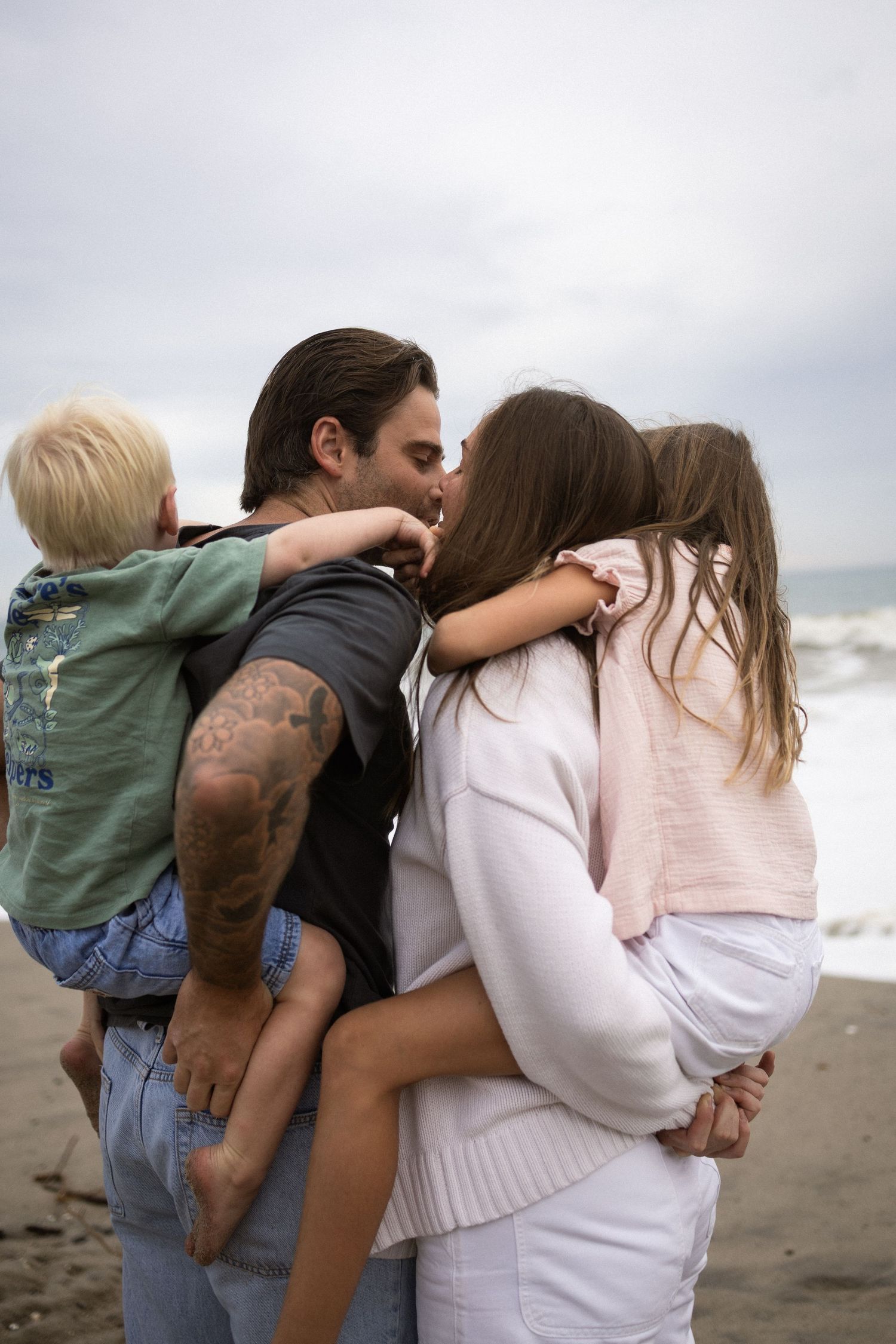 A tattooed man embraces a woman in white on a beach, with a toddler nearby and ocean waves in the background.