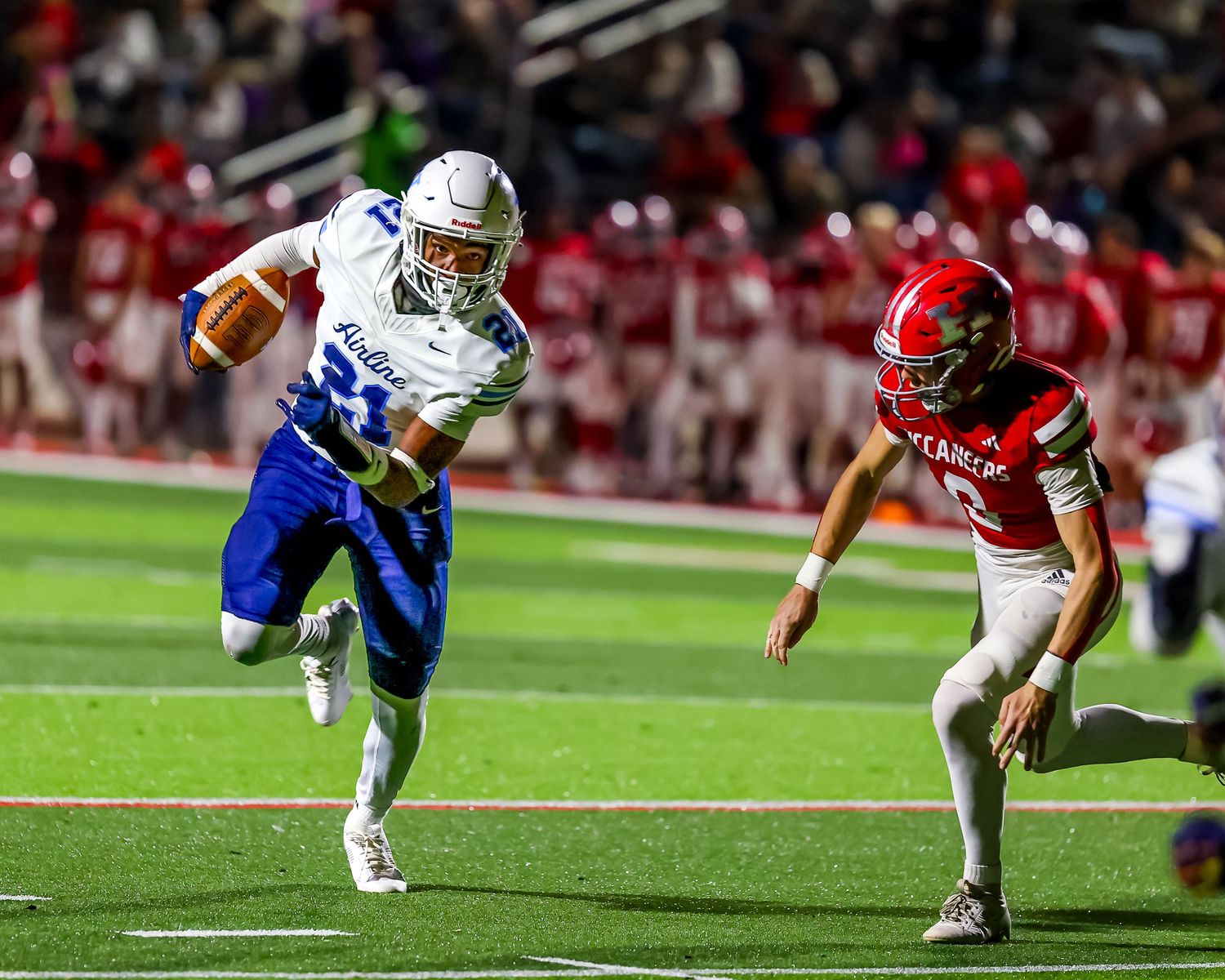 Running back in navy blue uniform carries football as defender in white jersey pursues during night game.