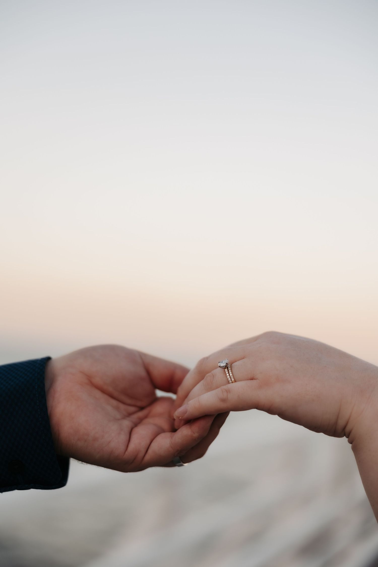Close-up of hands touching with engagement ring visible against soft sunset sky.