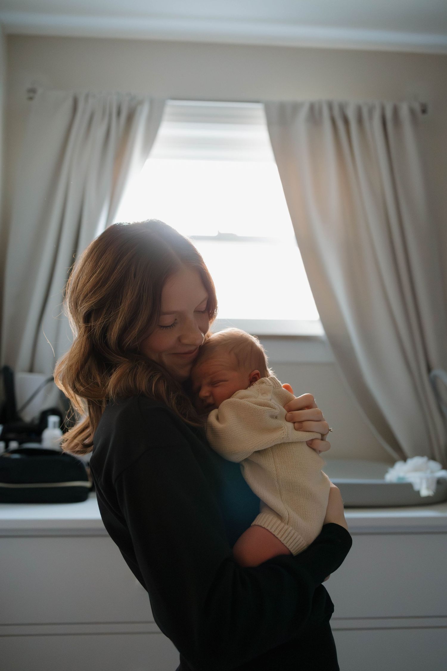 A tender moment between parent and newborn baby by a window with soft curtains in a sunlit room.