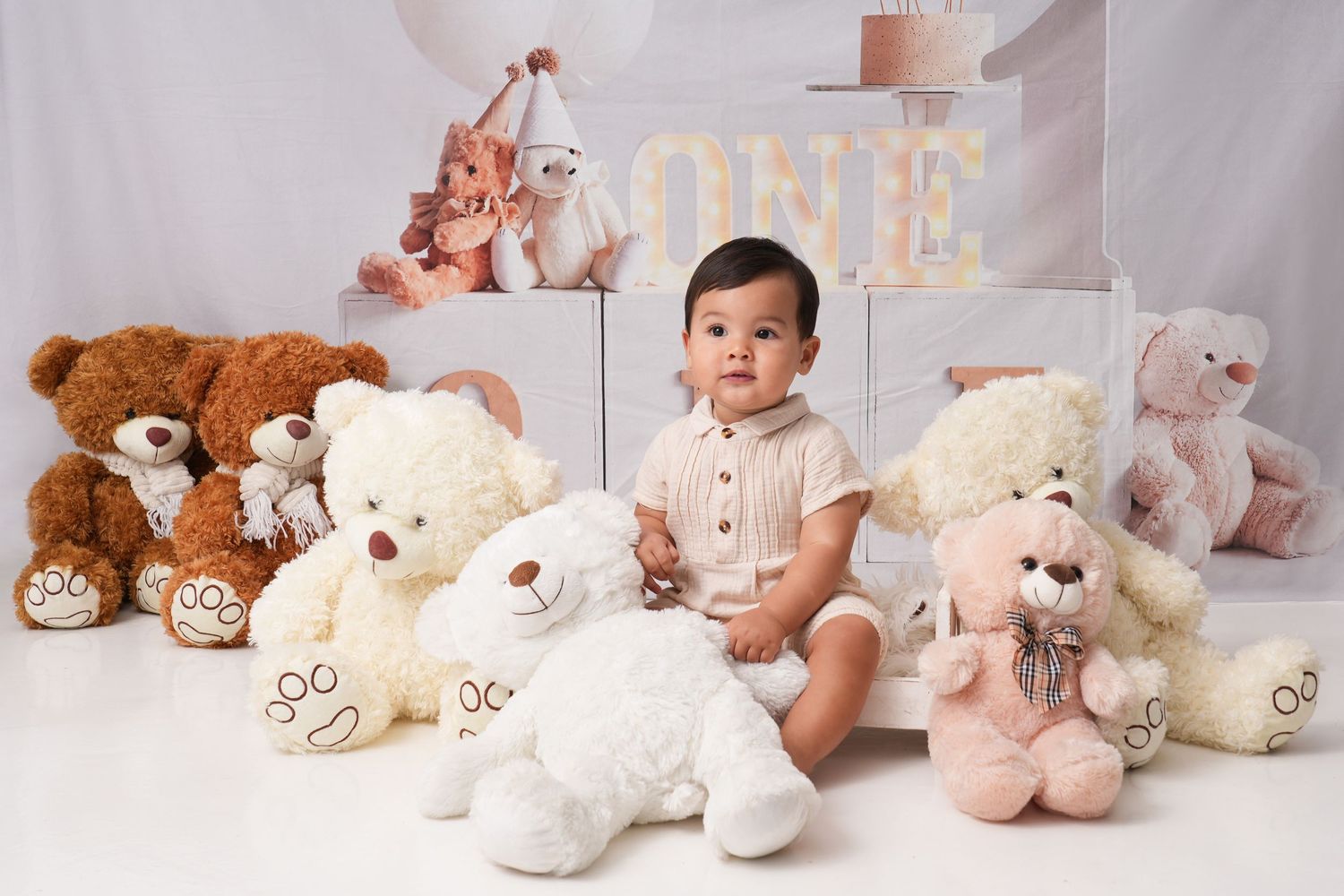 Baby in white outfit sits among soft pastel teddy bears and stuffed animals in studio setting.