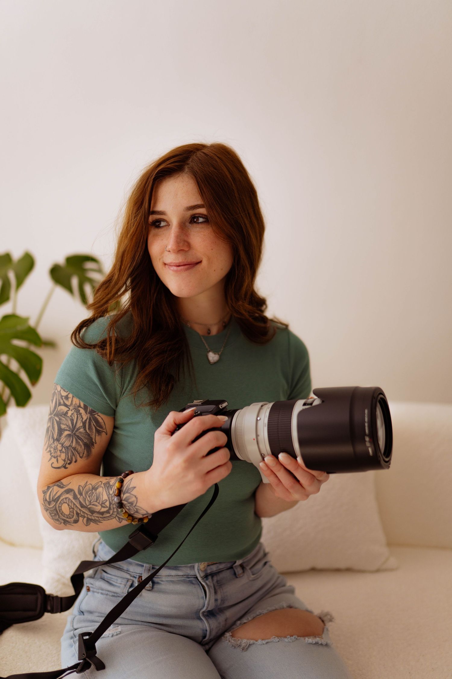 A photographer with tattoos holds a professional camera while sitting on a couch.