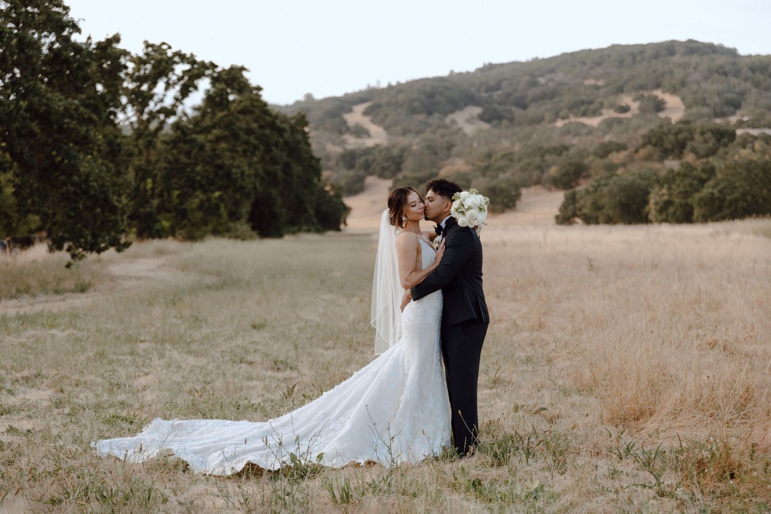 Couple in wedding attire standing together in a golden grass field with mountains in background.