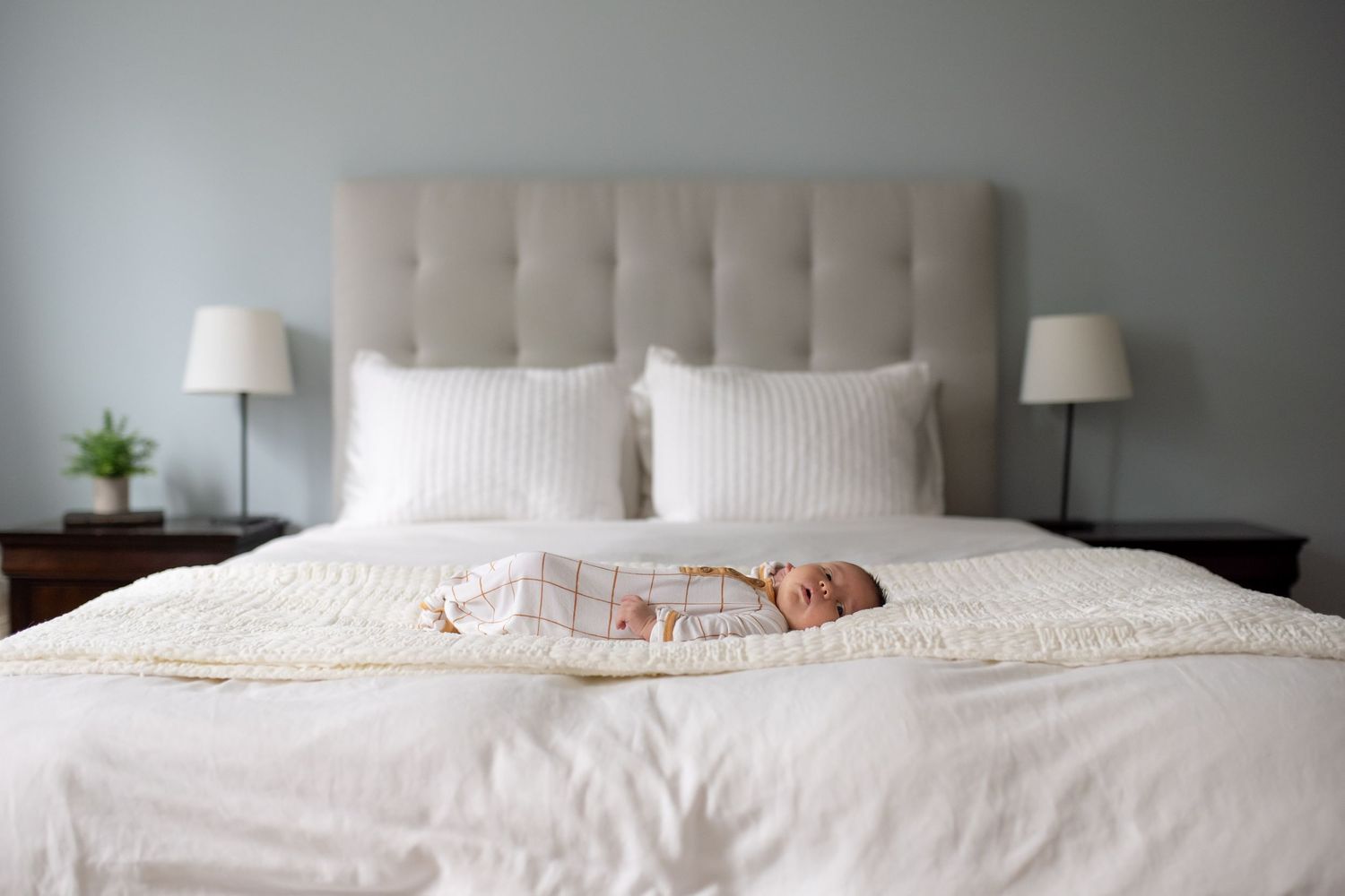A newborn baby sleeps peacefully on a plush white bed with tufted gray headboard and matching bedside lamps.