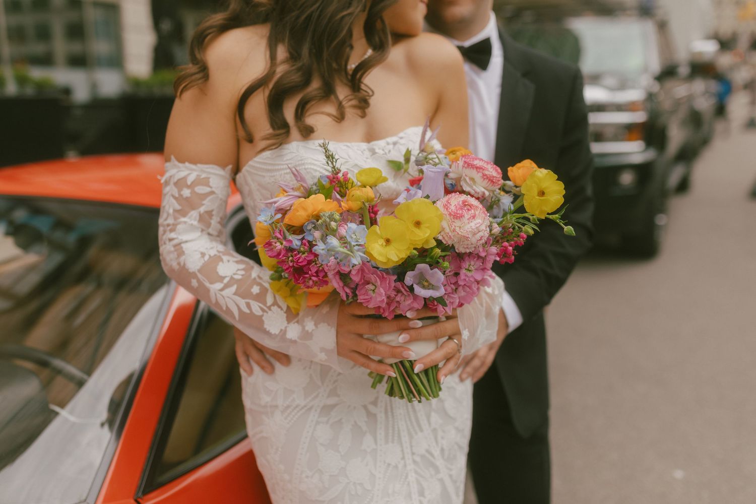 A wedding bouquet of pink and yellow flowers held by someone in a white lace wedding dress standing next to a red car.