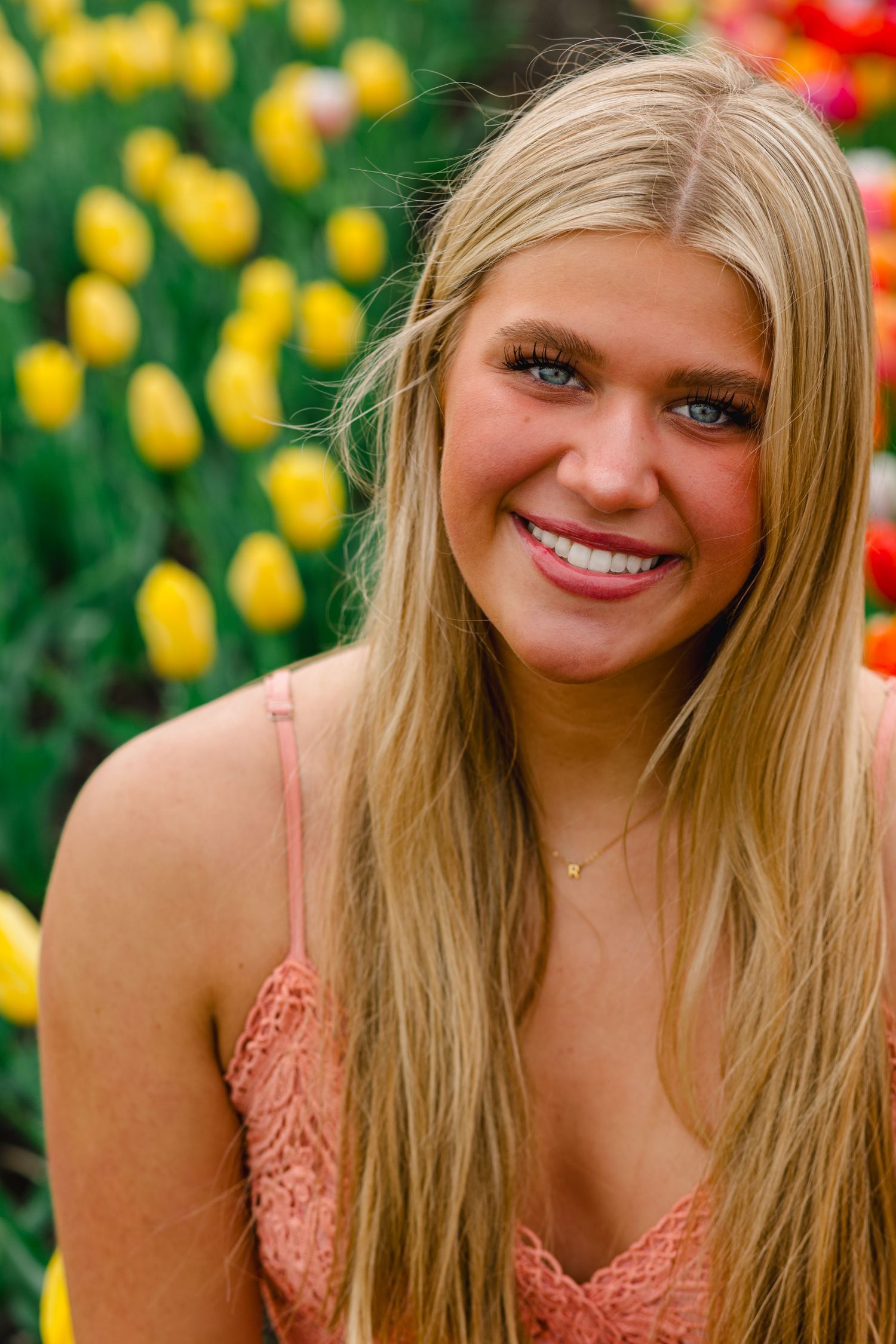 Smiling woman with blonde hair poses outdoors against colorful yellow tulips in spring garden setting.