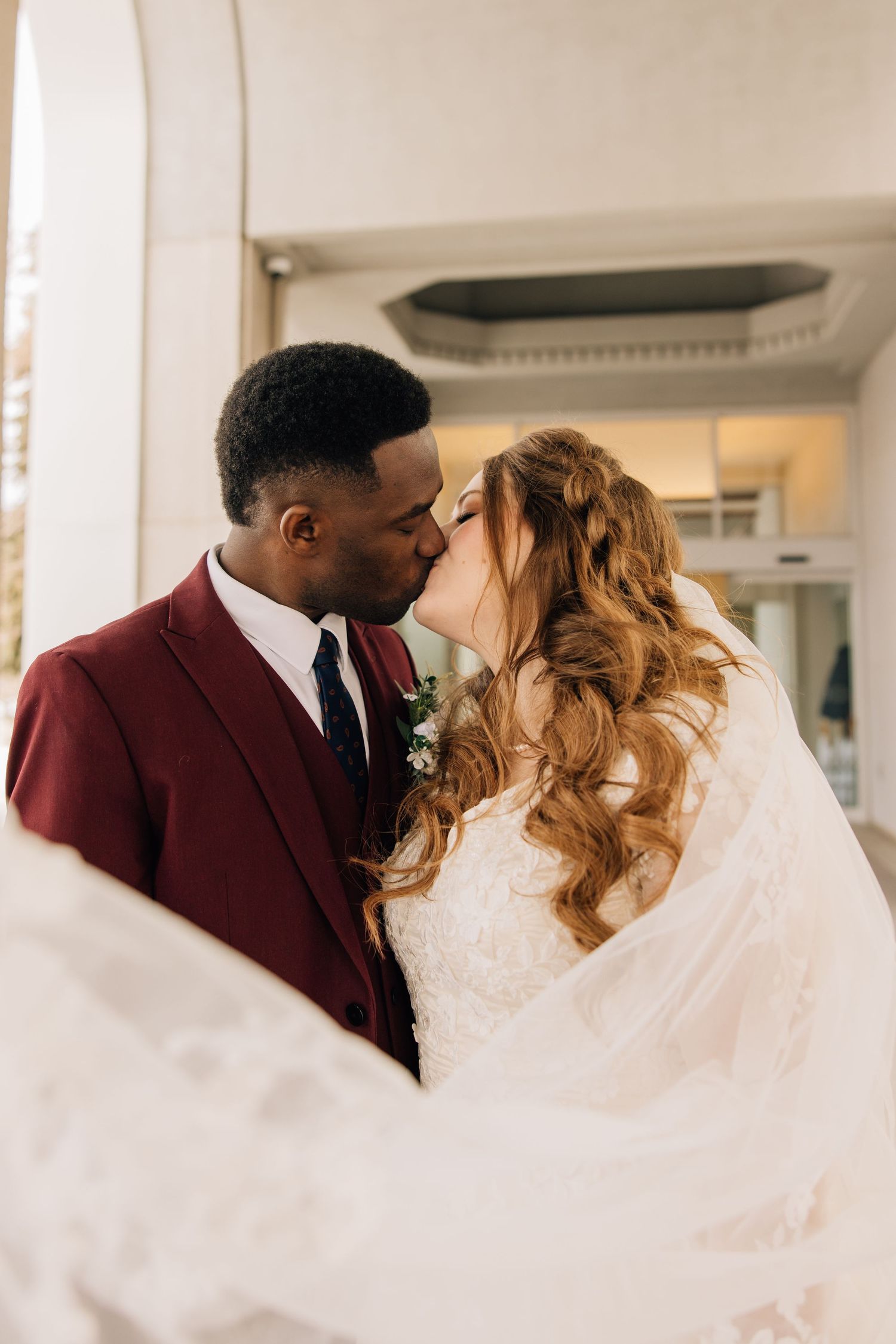 A romantic wedding photo of a couple sharing a kiss, with the bride in white and groom in burgundy suit.