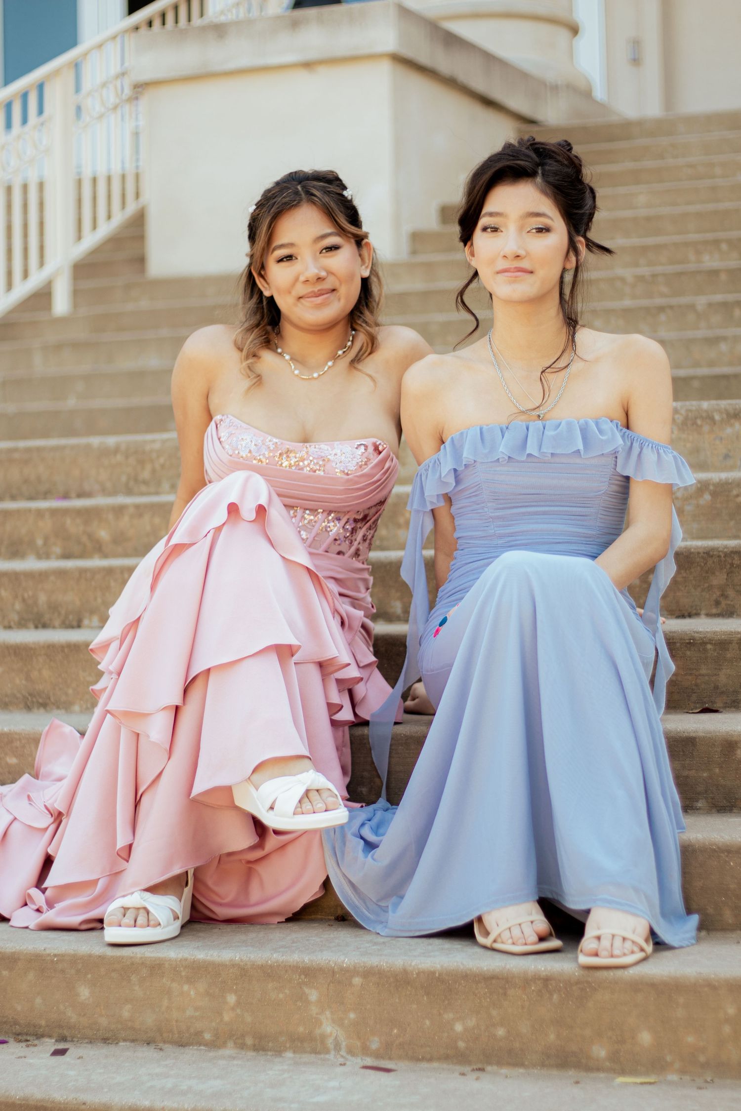 Two elegant formal dresses, one in dusty rose with ruffles and one in silver gray with off-shoulder design in a sunlit hallway.