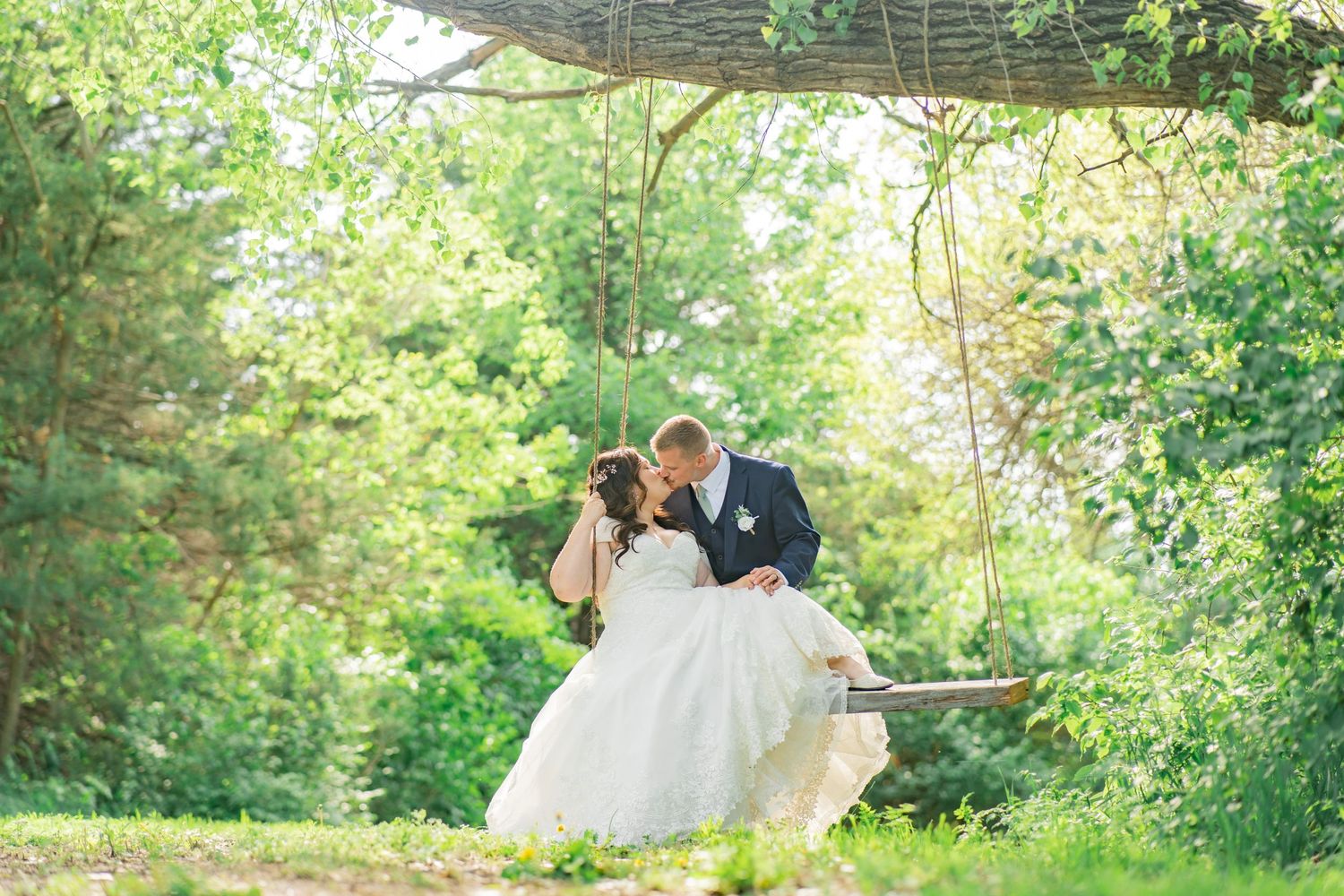 Bride and groom sharing a romantic kiss on a wooden swing surrounded by lush green trees outdoors.