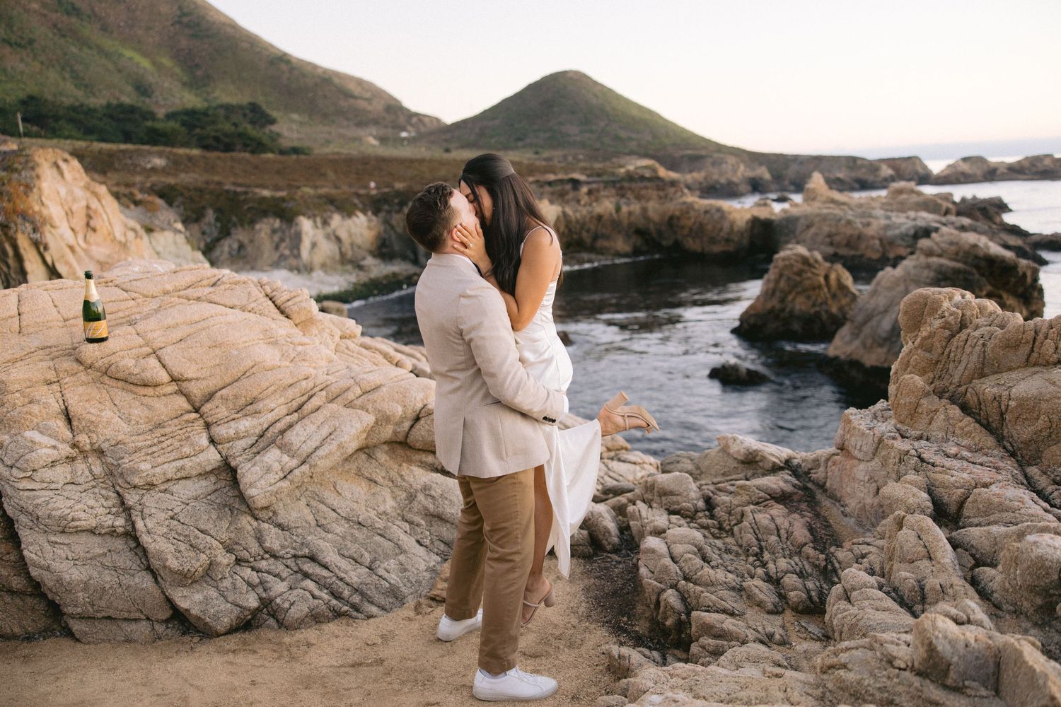 A couple embraces on rugged coastal rocks at sunset along the California coastline.