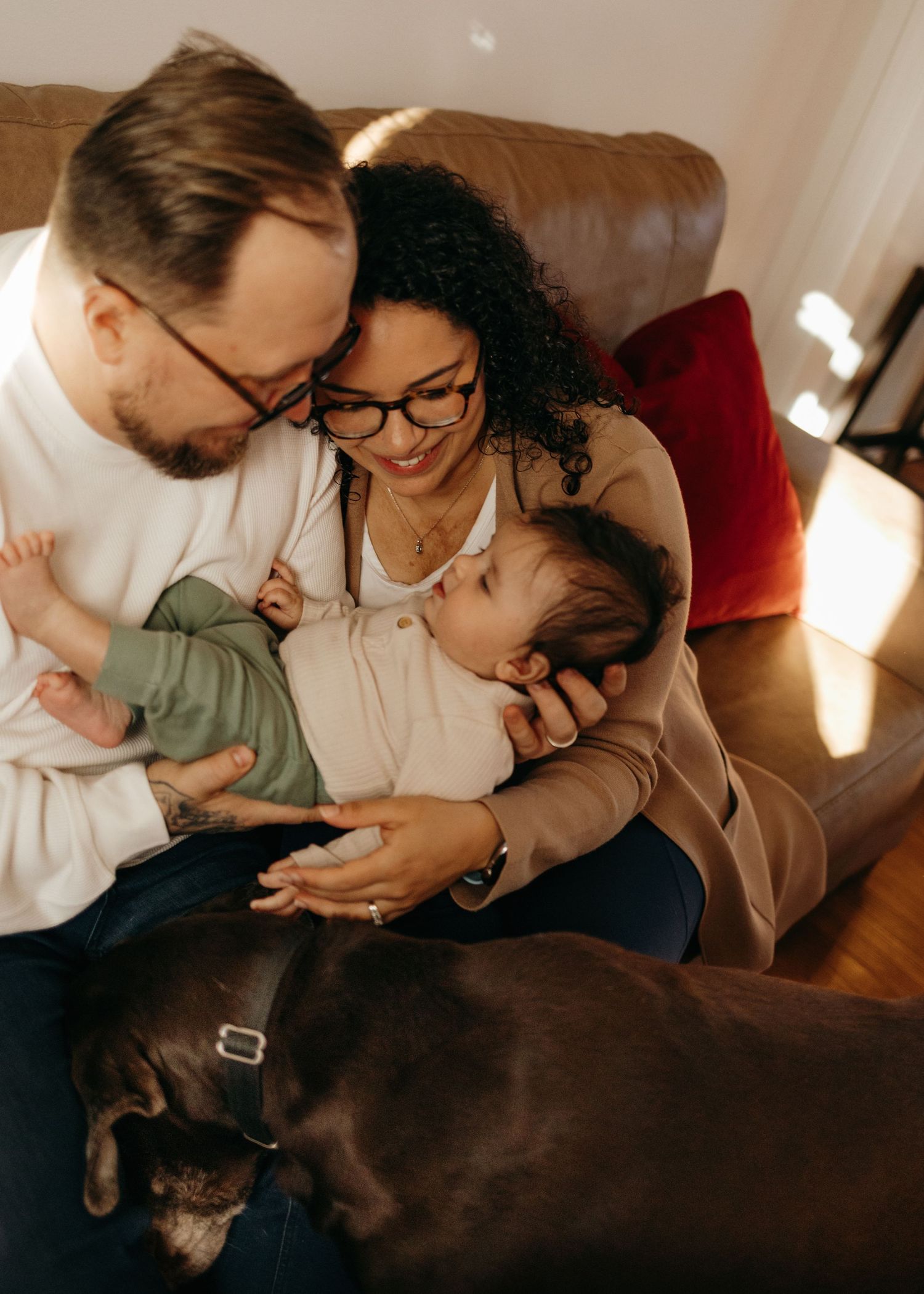 A family cuddles together on a brown leather couch while a dark dog lounges nearby in warm sunlight.