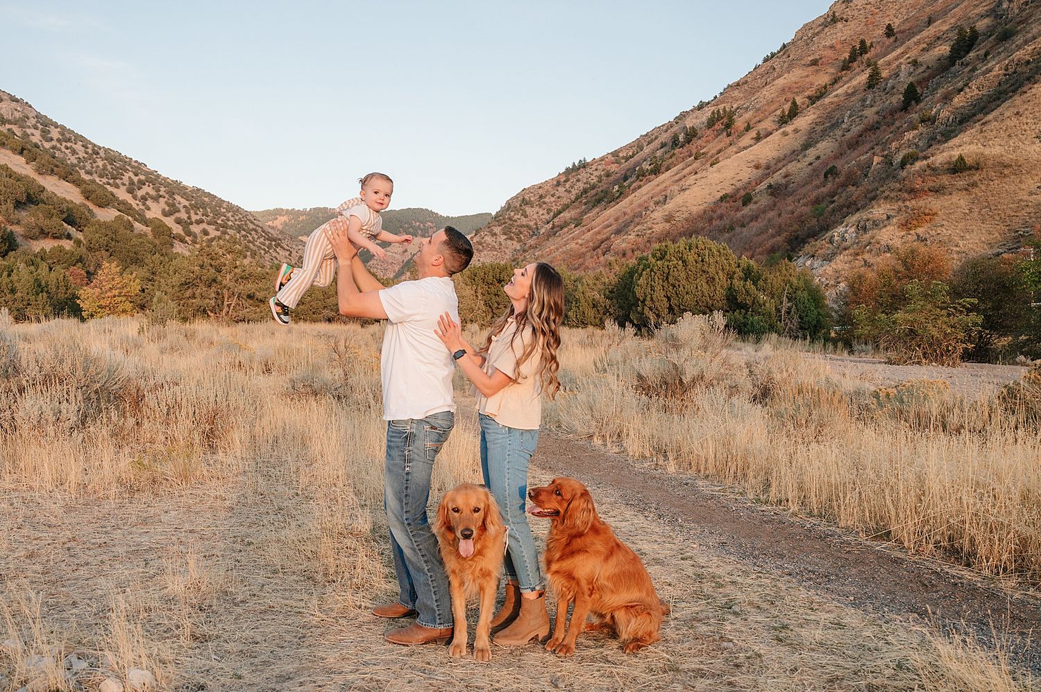 A couple stands with their two dogs against a backdrop of rolling hills and golden grass.