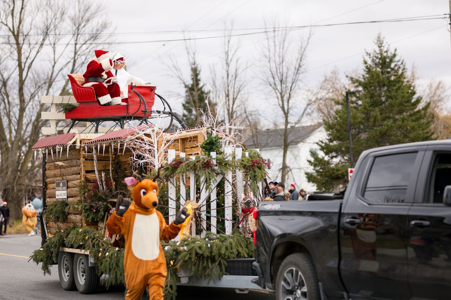 A holiday parade float featuring Santa and a reindeer character beside evergreen decorations and a black pickup truck.