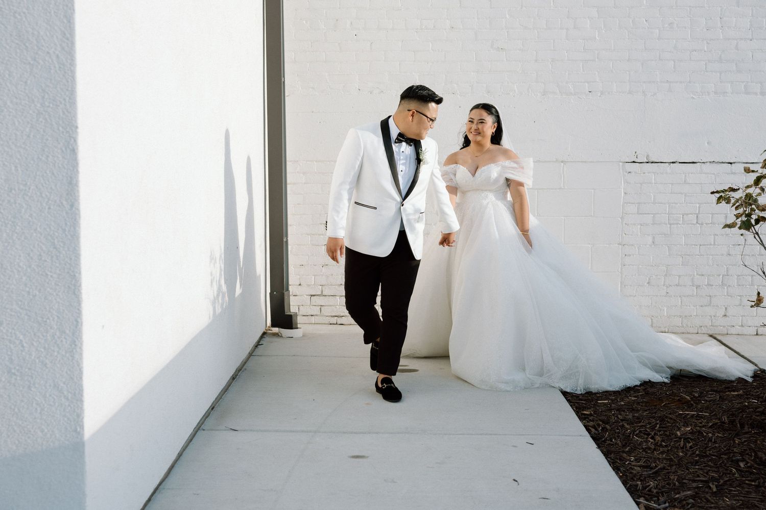 A wedding couple walking together against a white wall in an outdoor setting.