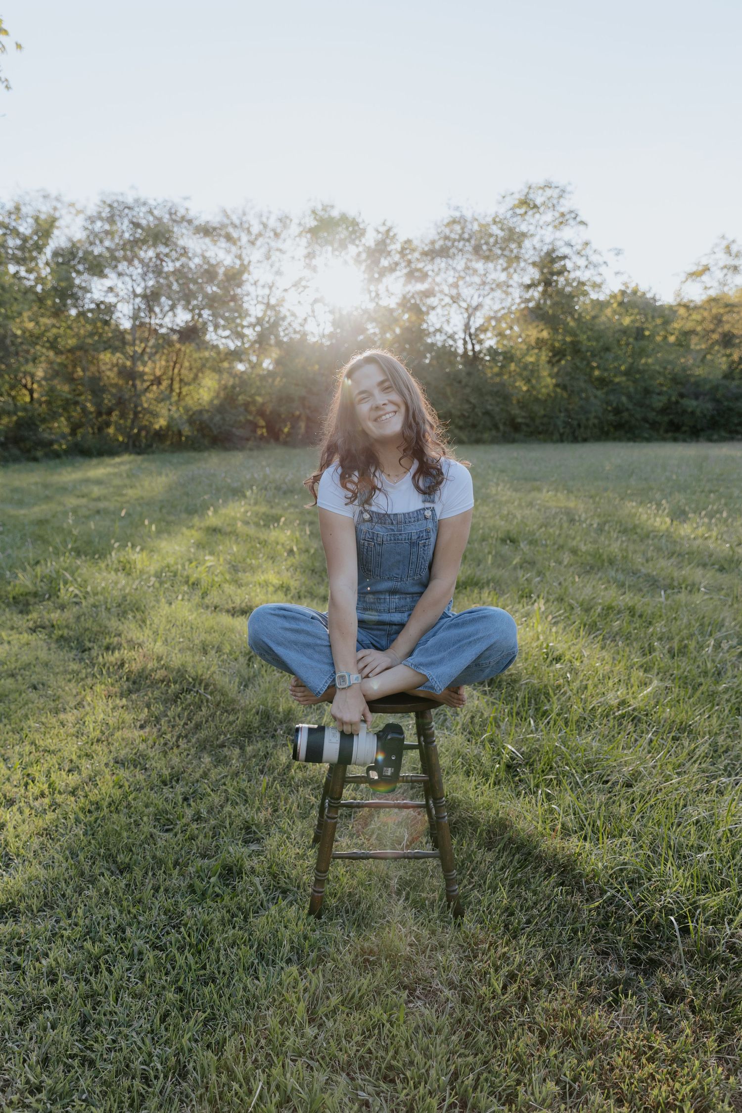 Someone in denim overalls sits casually on a wooden stool in a sunlit grassy field with a camera.