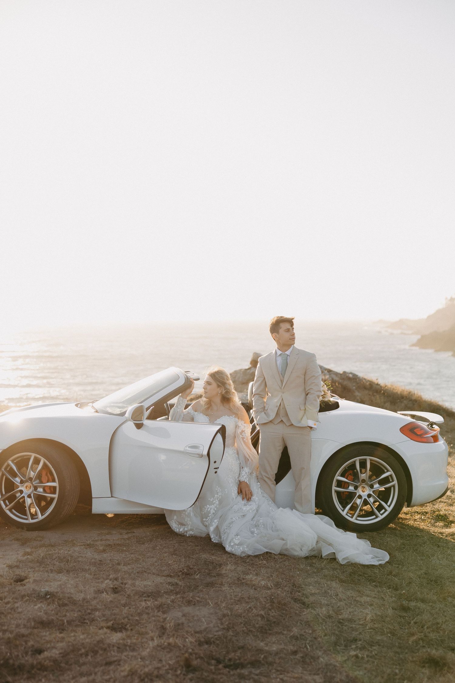 A couple in wedding attire poses with a white luxury convertible sports car on a scenic coastal cliff at sunset.