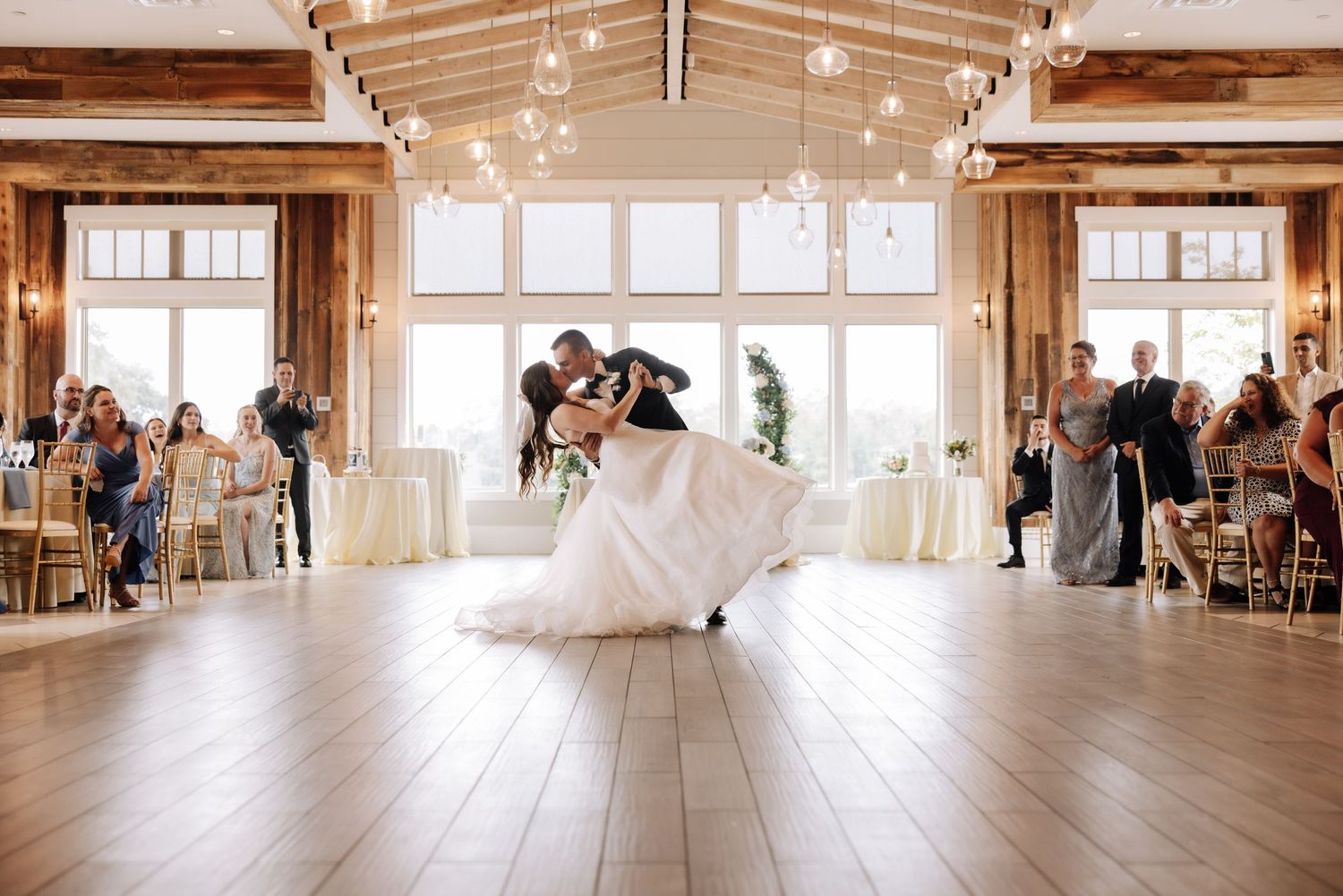 A graceful dance between two people in wedding attire inside a rustic venue with exposed beams and string lights.
