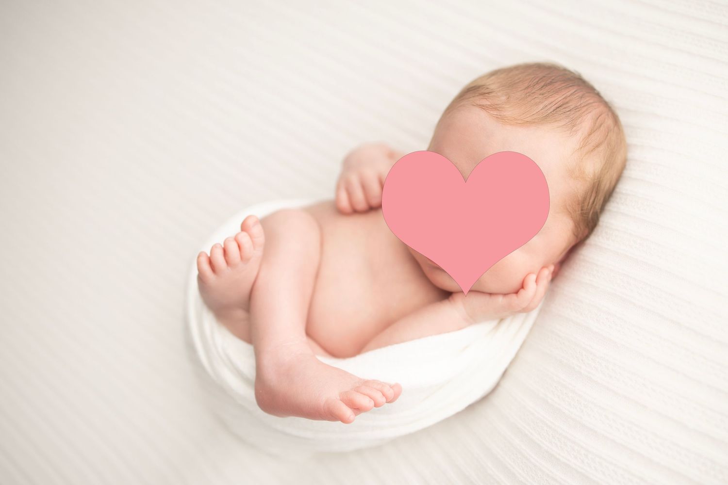 Newborn baby photographed on white fabric with pink heart covering face.