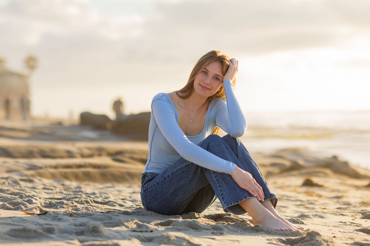 Person in blue sweater and jeans sitting on sandy beach at sunset.