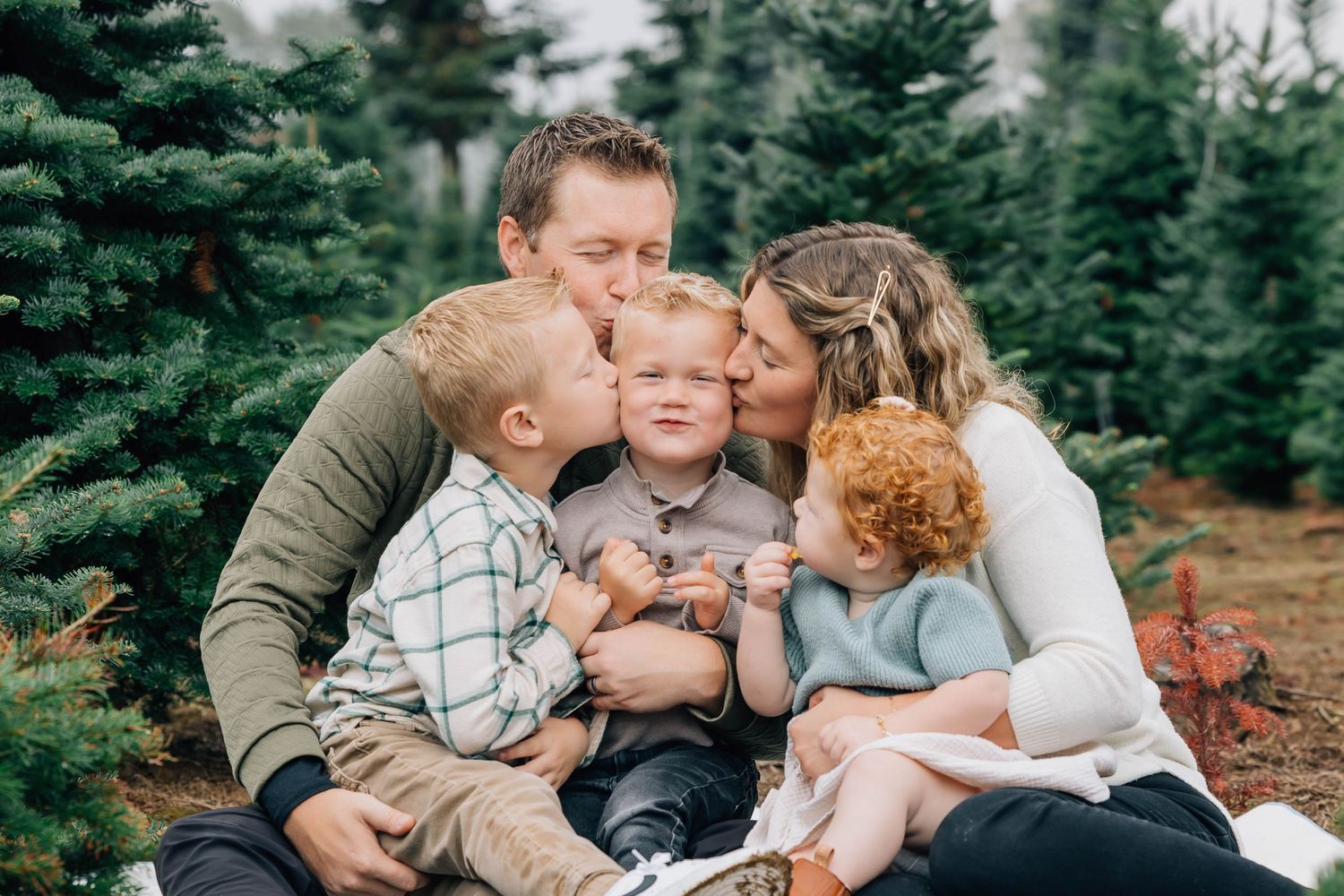 Happy family with three young children sharing kisses at a Christmas tree farm.