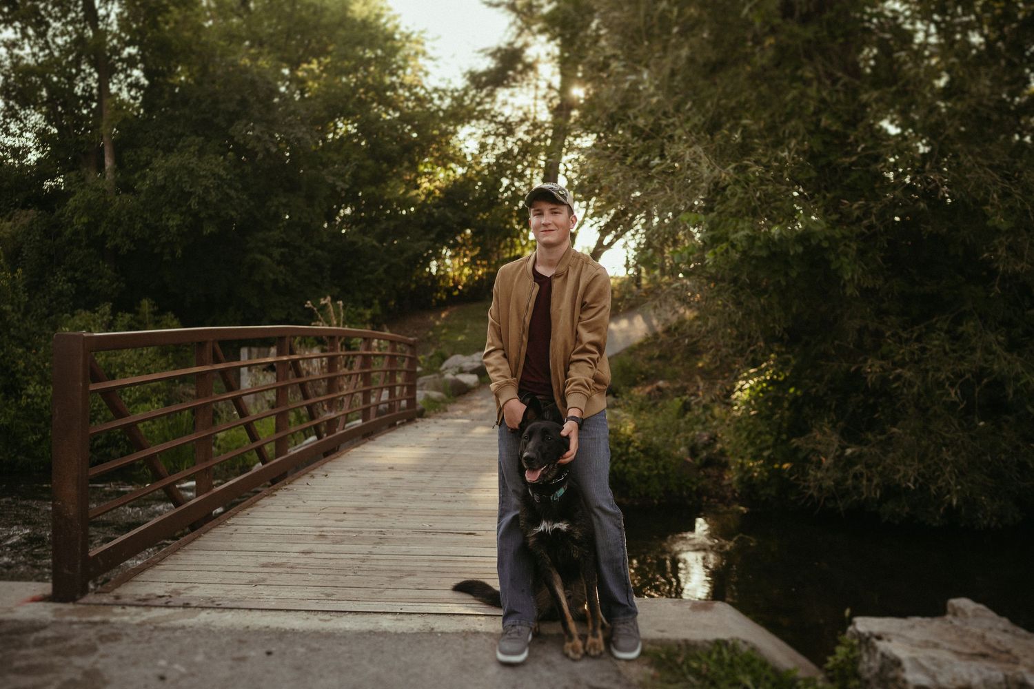 A person in a brown jacket stands with their dog on a wooden bridge during golden hour sunset.