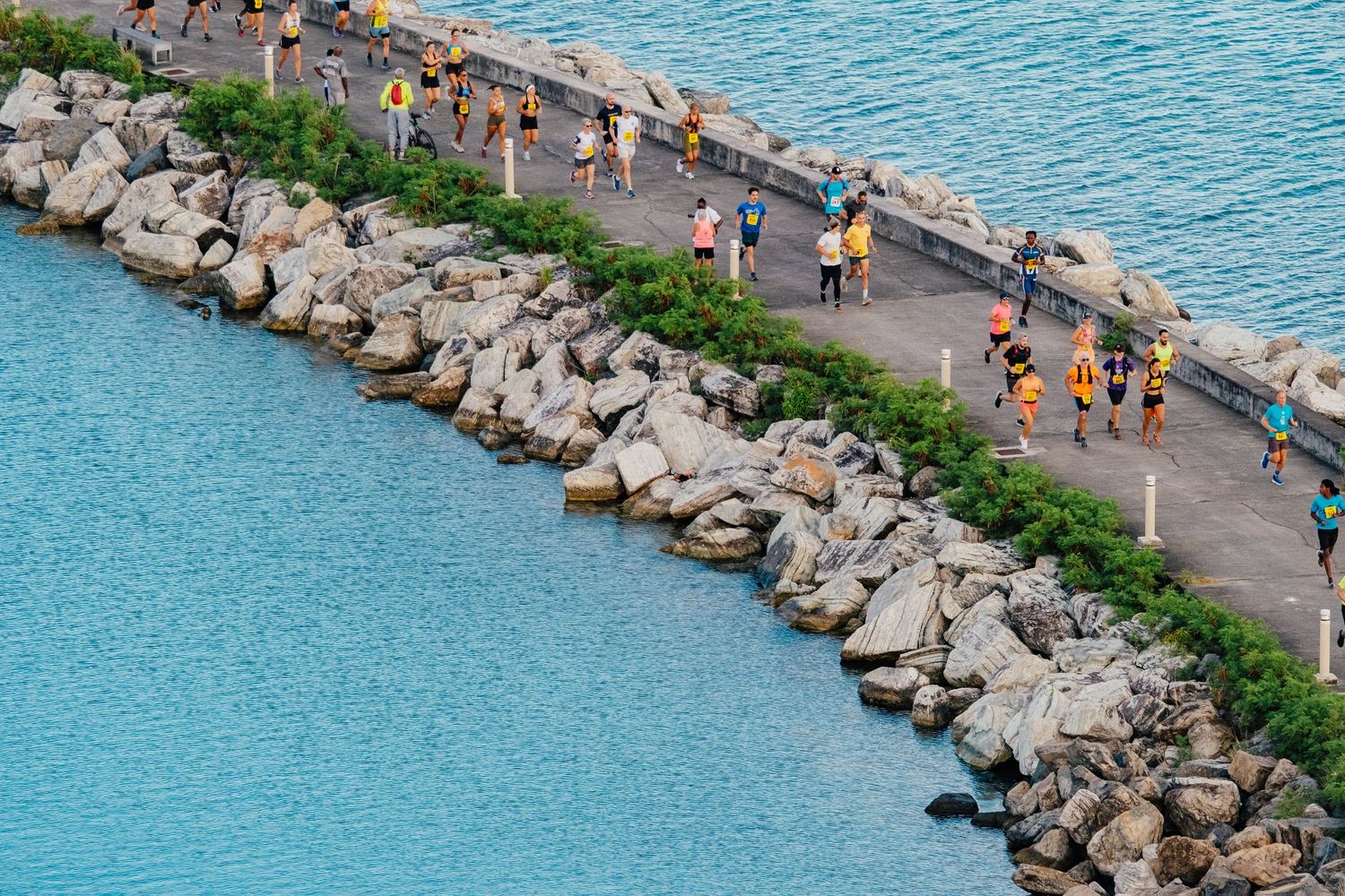 Aerial view of runners exercising on a curved stone jetty path surrounded by bright blue water.