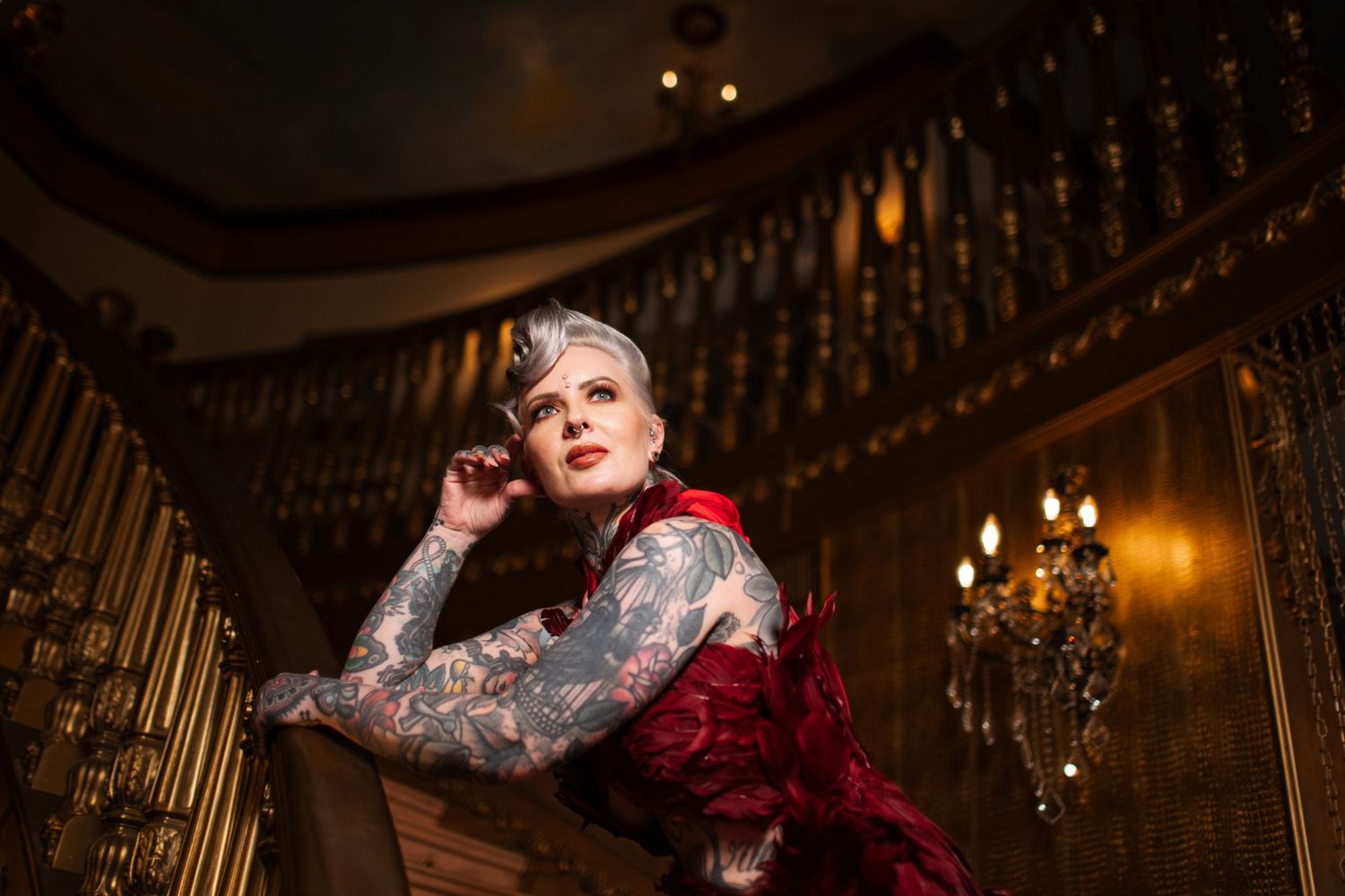 Figure in vintage red velvet dress poses near ornate staircase with crystal chandelier in elegant interior setting.