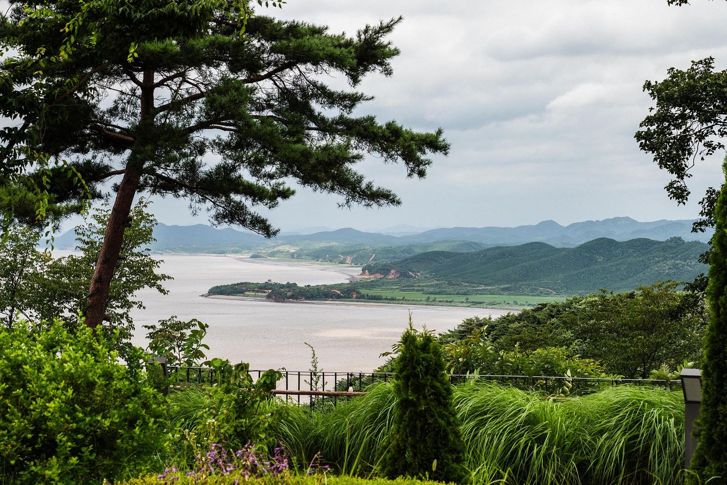 Scenic overlook of a tranquil bay with green hills, lush trees, and a distant red ship under cloudy skies.