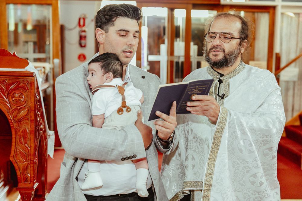 A man holds a baby during a baptism ceremony while a priest reads from a religious book in a church.