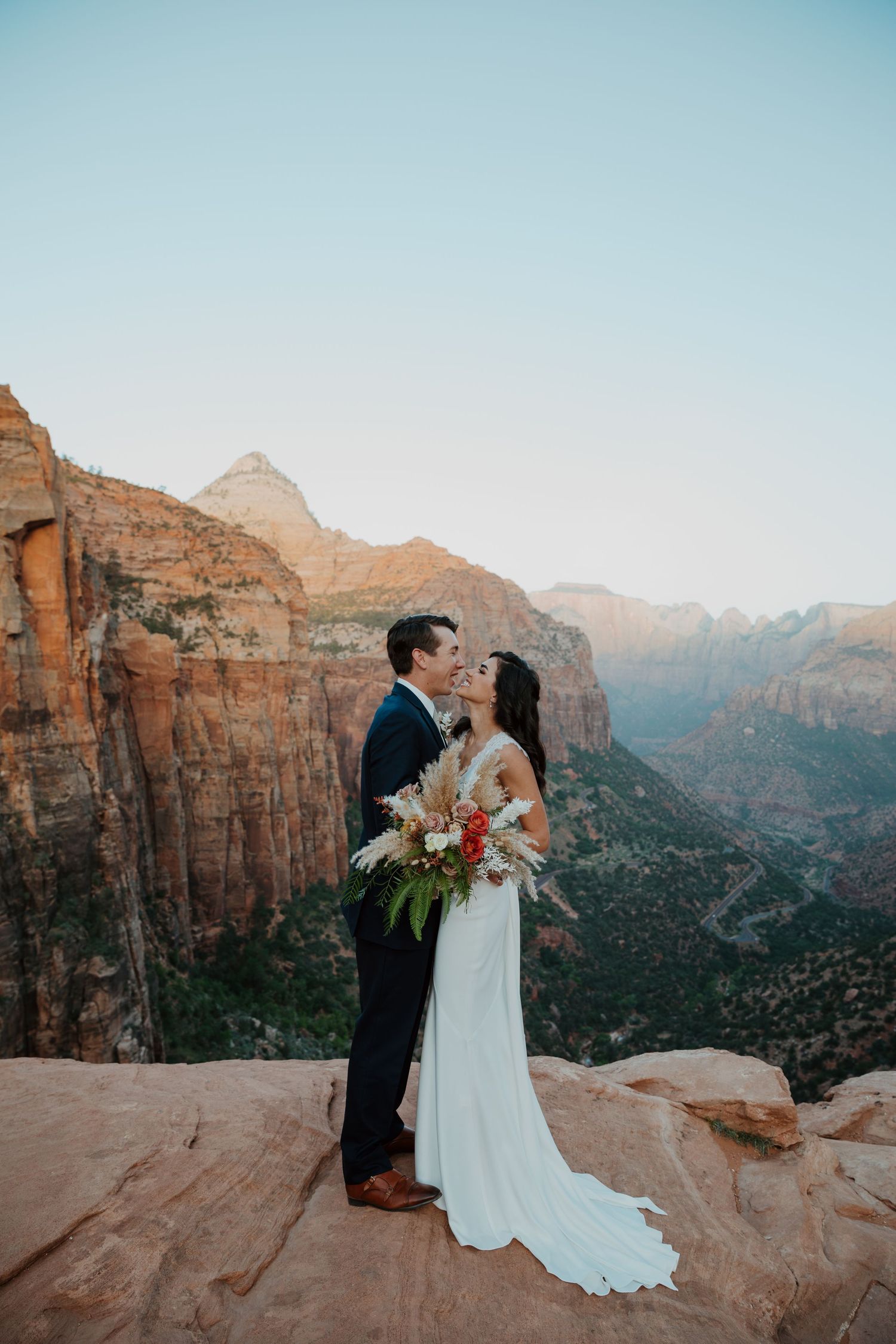 Bride and groom share a romantic kiss on a cliff overlooking Zion National Park canyon at sunset.