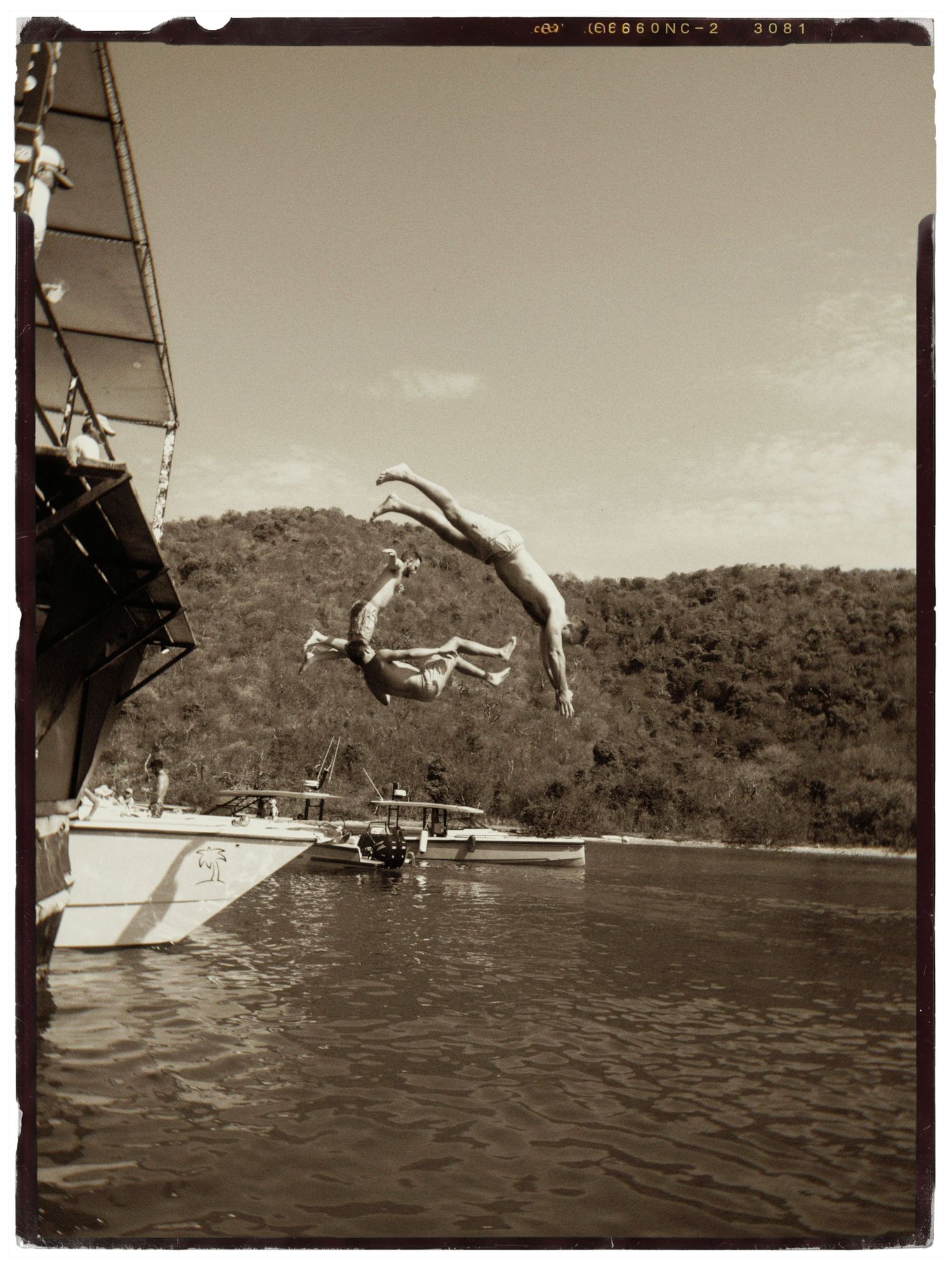 Two people diving off a sailboat into calm water with tree-covered hills in the background, captured in black and white.